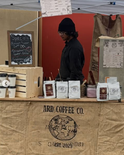 A young man wearing a black beanie and glasses working at a coffee stand with a burlap table cover printed with 'ARD COFFEE CO.' and a globe logo. There are coffee bags, paper cups, and a chalkboard sign listing treats like brown sugar lattes and strawberry steak matcha.