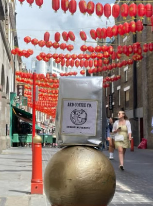 A street decorated with many red lanterns hanging overhead, with a large golden sphere on the sidewalk, and a coffee bag labeled 'ARAD COFFEE CO. Ethiopia.' Passing pedestrians are visible in the background.