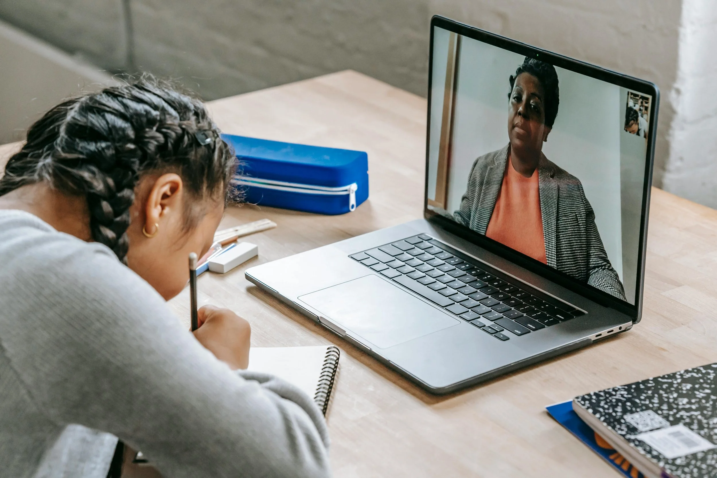 Person sitting at a desk, taking notes while on a video call on a laptop with another woman.