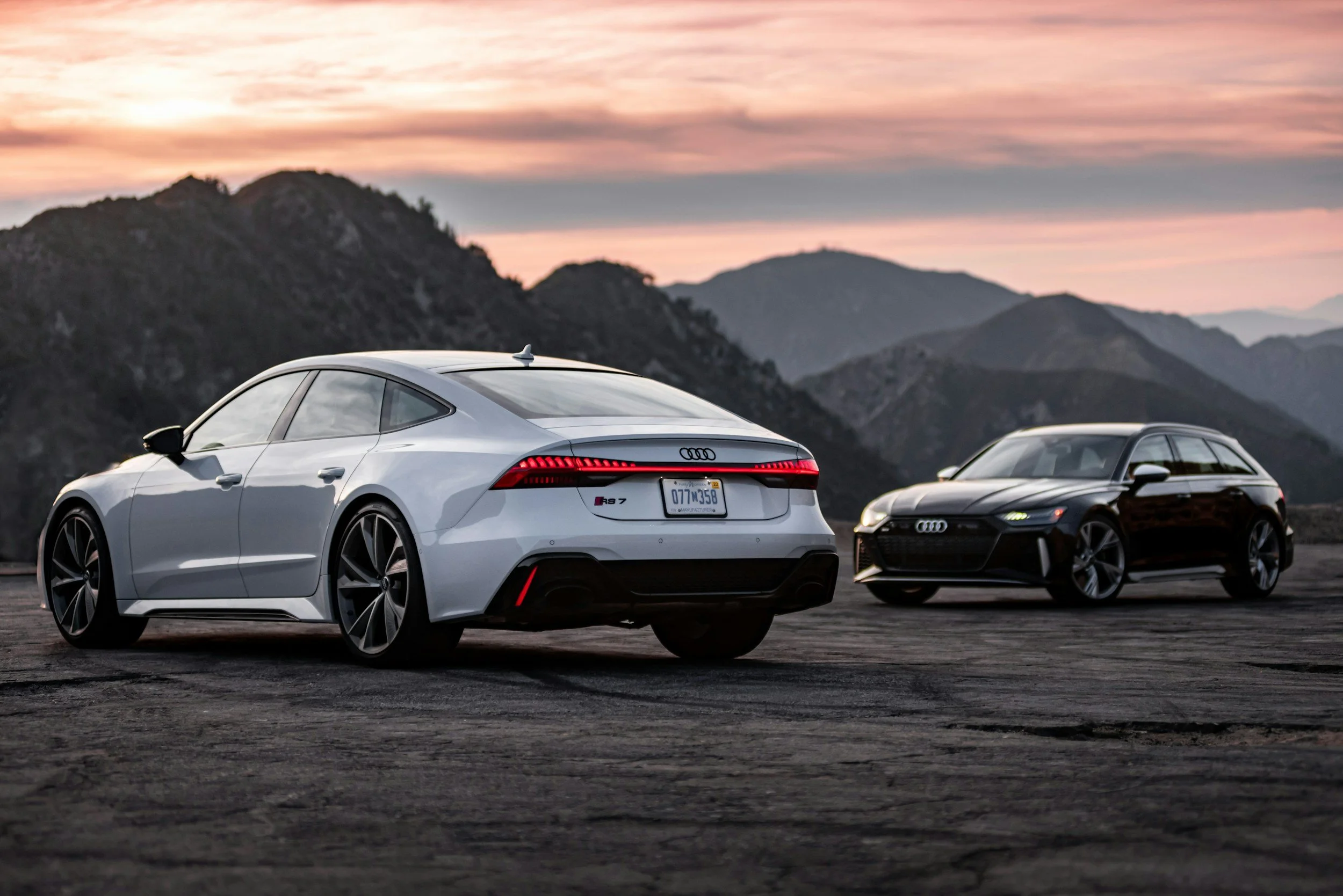 Two Audi cars, a white RS 7 and a black station wagon, parked on an open mountain landscape during sunset.