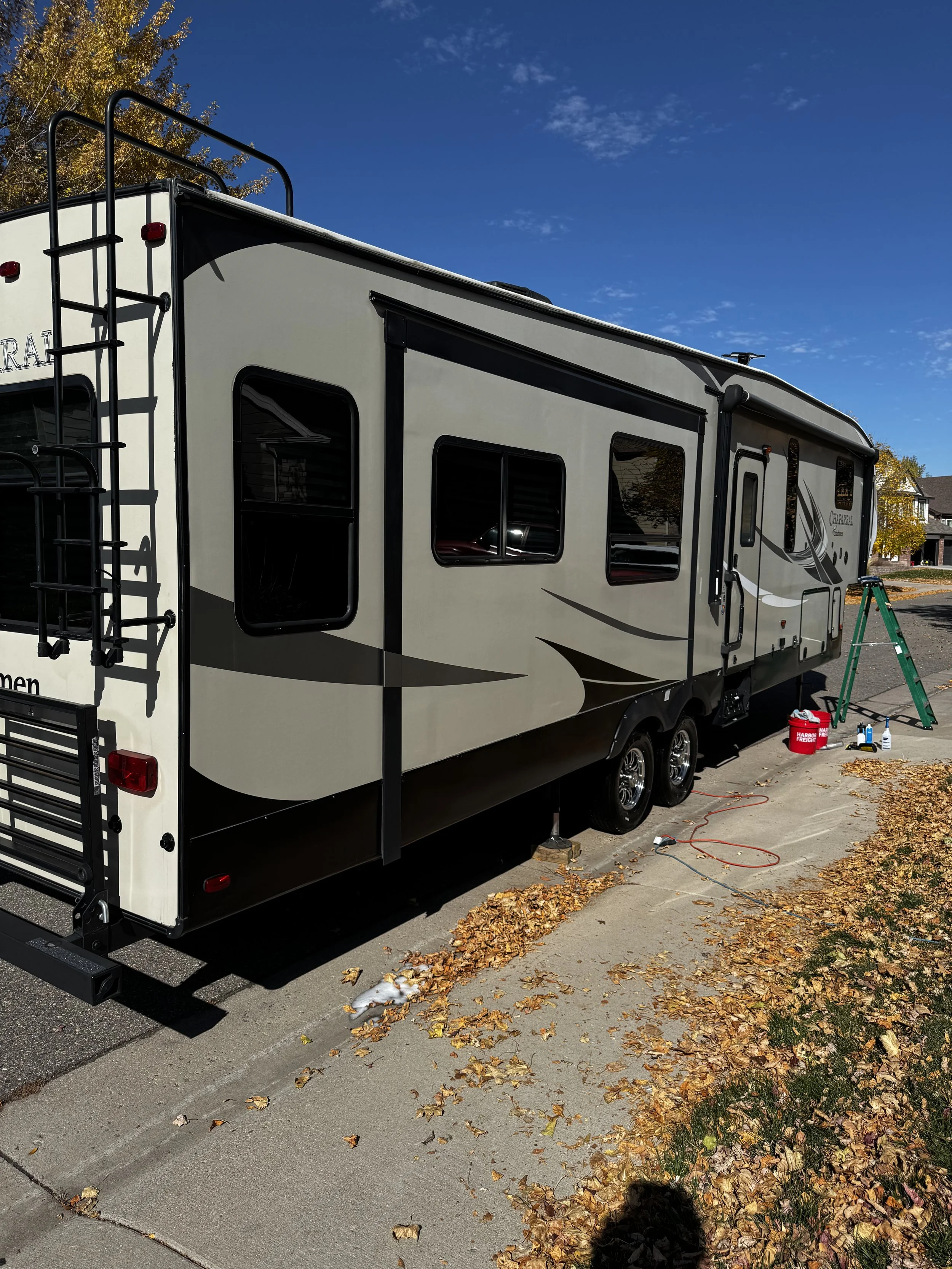 A large beige and black RV parked on the side of a street with fallen autumn leaves. A green ladder and cleaning supplies are nearby.