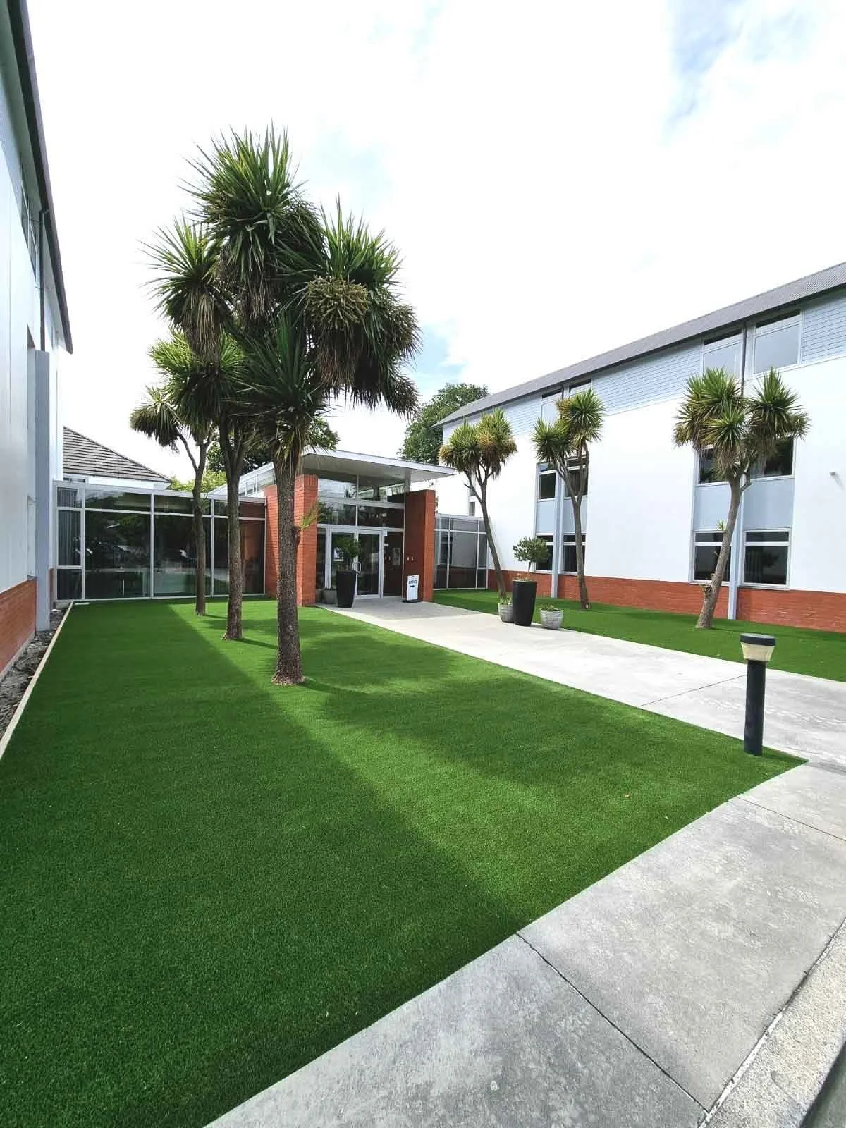 Trimmed artificial grass lawn lined with tall palm trees along a walkway outside a modern commercial building with brick and white walls.