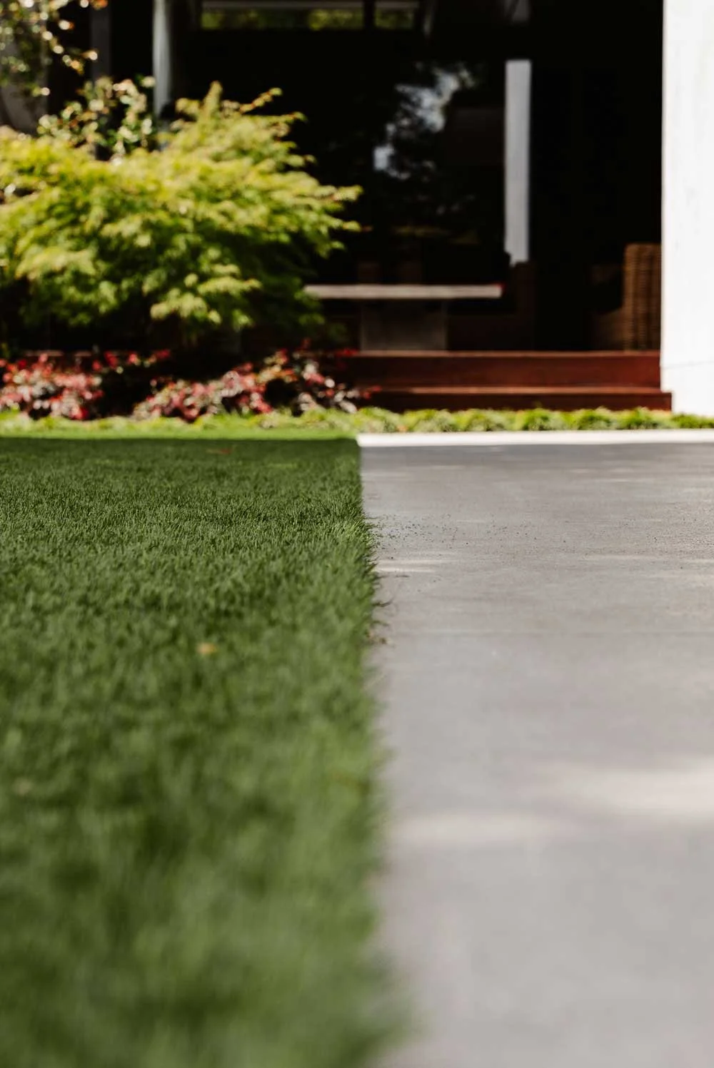 Close-up view of a garden with artificial grass on the left and a concrete pathway on the right, leading to a house with a porch surrounded by greenery.