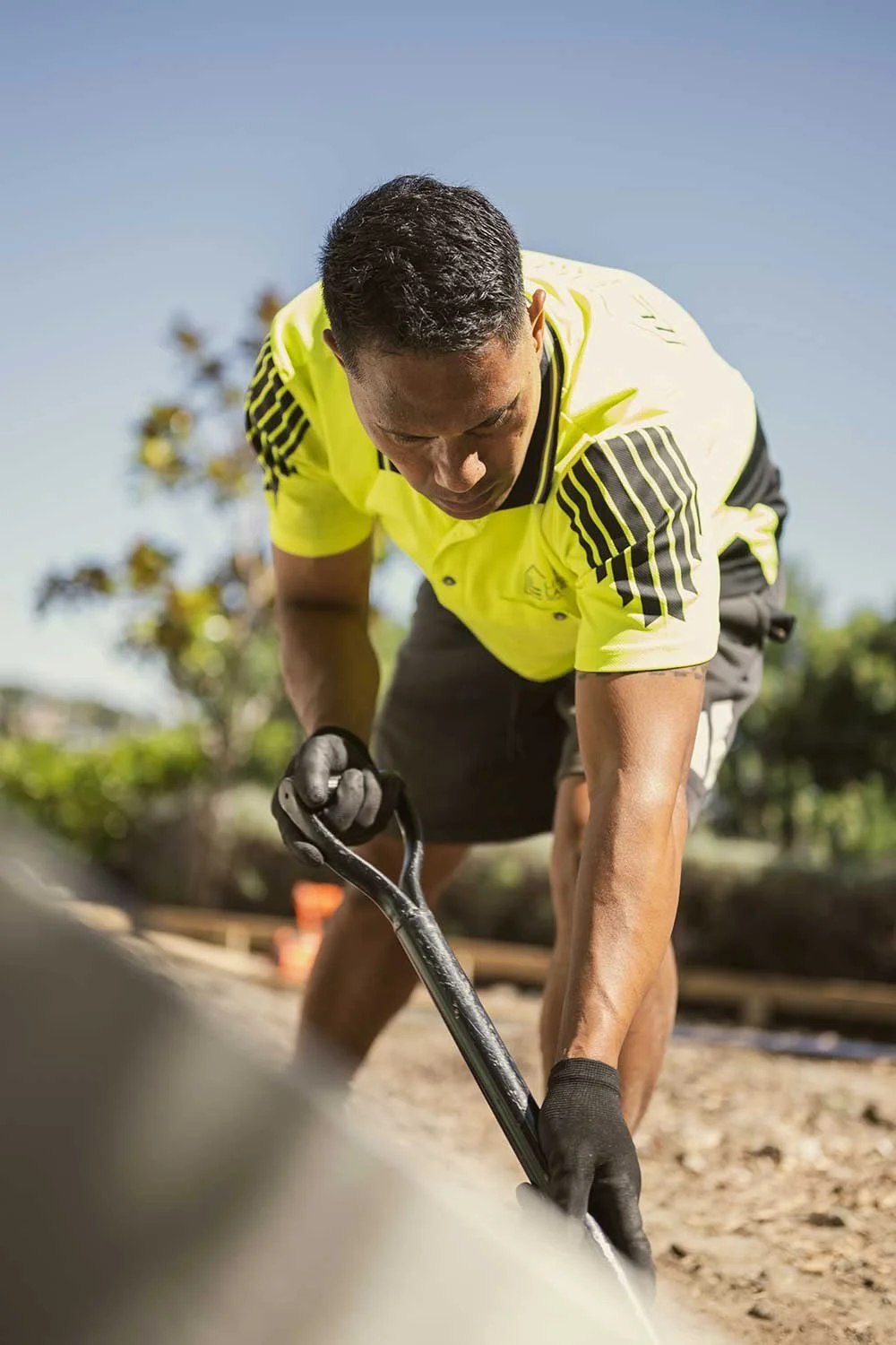 A man wearing a yellow shirt and gloves is working outdoors with a tool on a construction site.