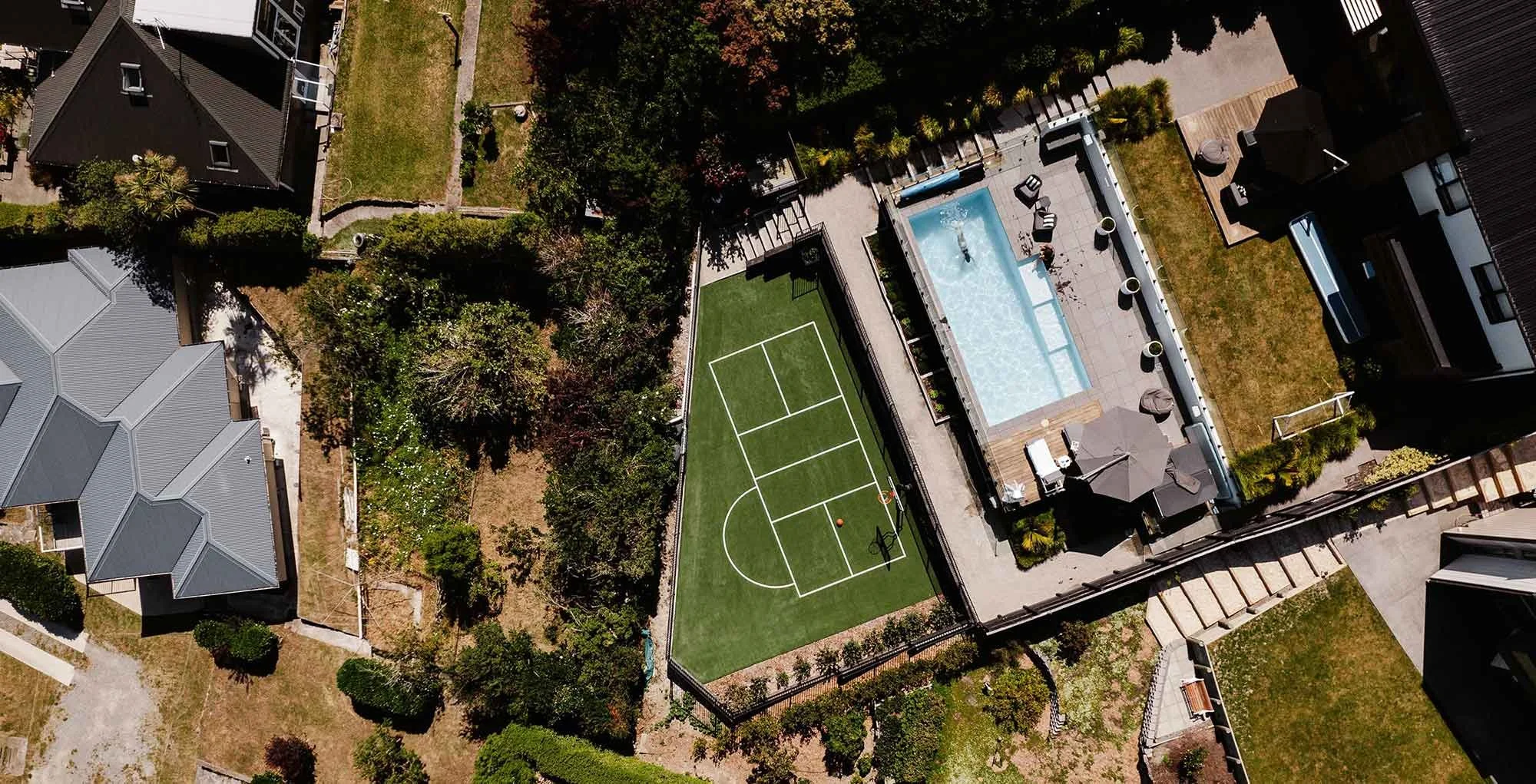 An aerial view of a backyard featuring a basketball court, a swimming pool, and surrounding greenery, with neighbouring houses visible.