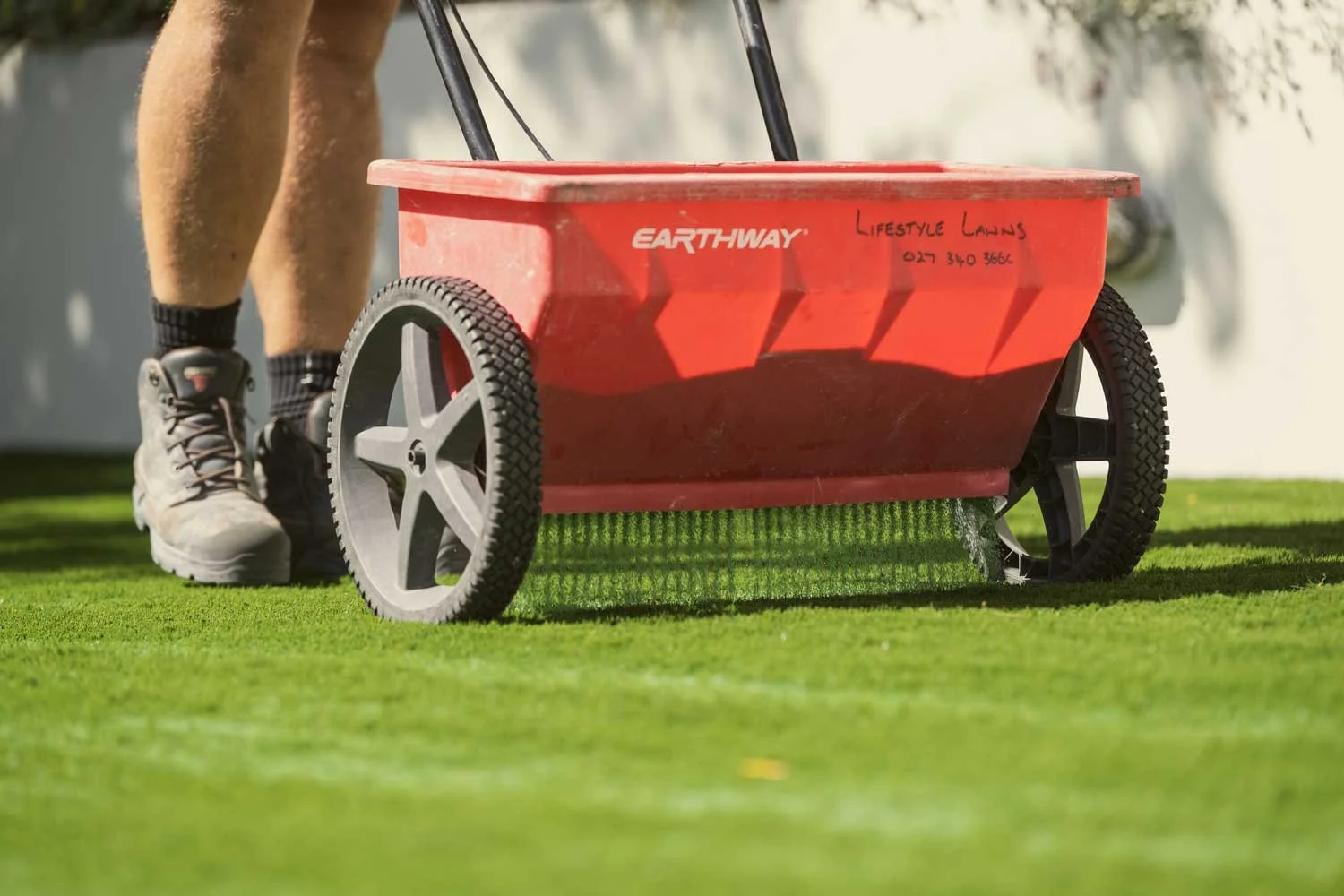 Person pushing a red wheelbarrow with infill onto artificial grass underneath.