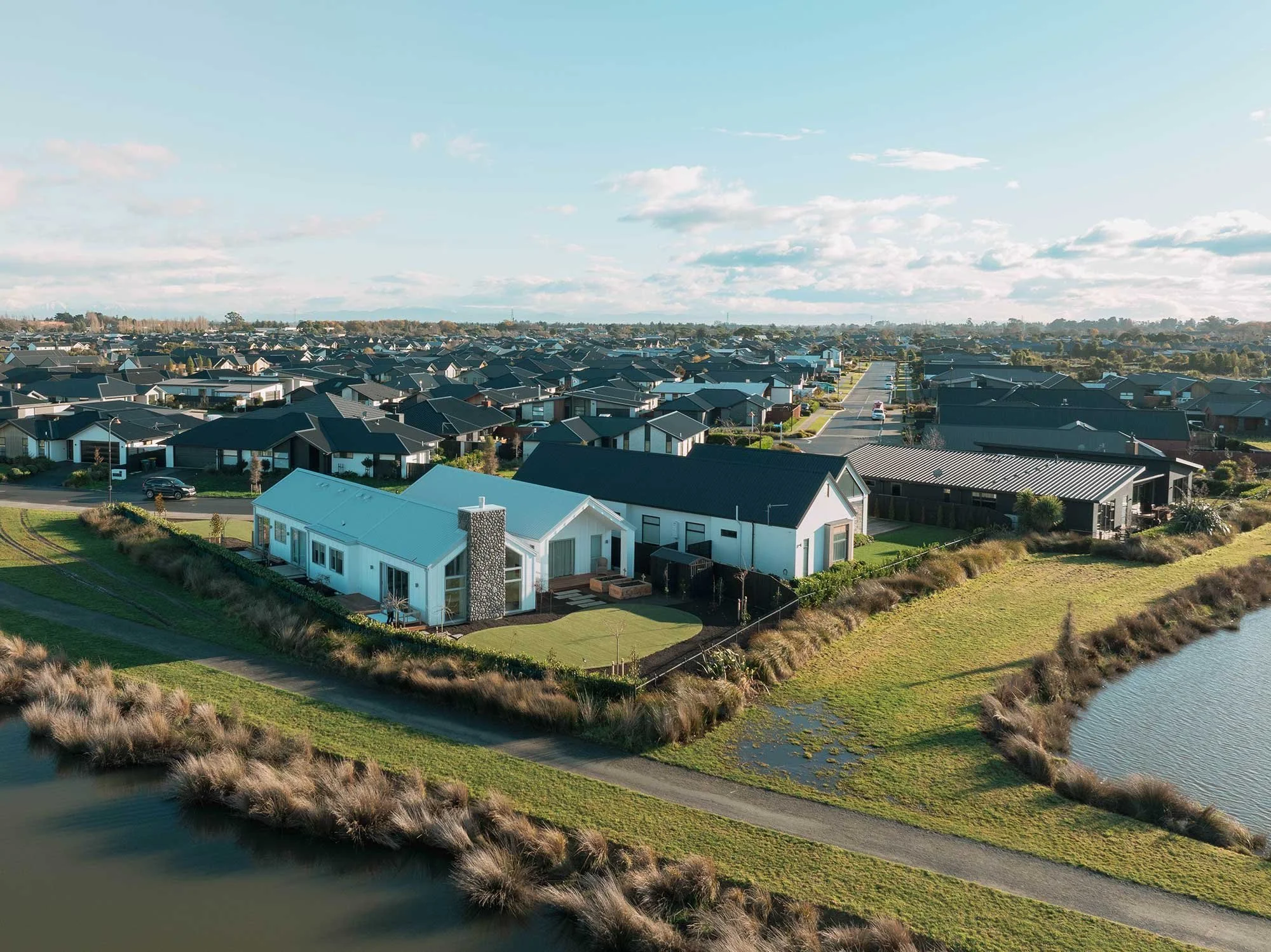 Aerial view of modern suburban homes beside a pond, landscaped with green lawns and surrounded by open space under a clear sky.