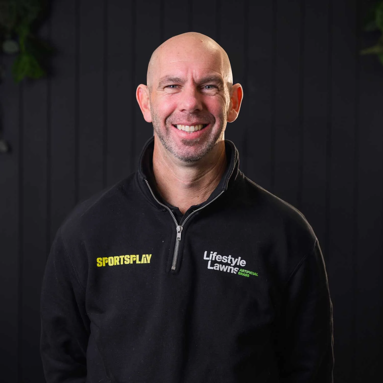 Damian Vercoe, Business Manager at Lifestyle Lawns, smiling, wearing black jacket and standing against black background.