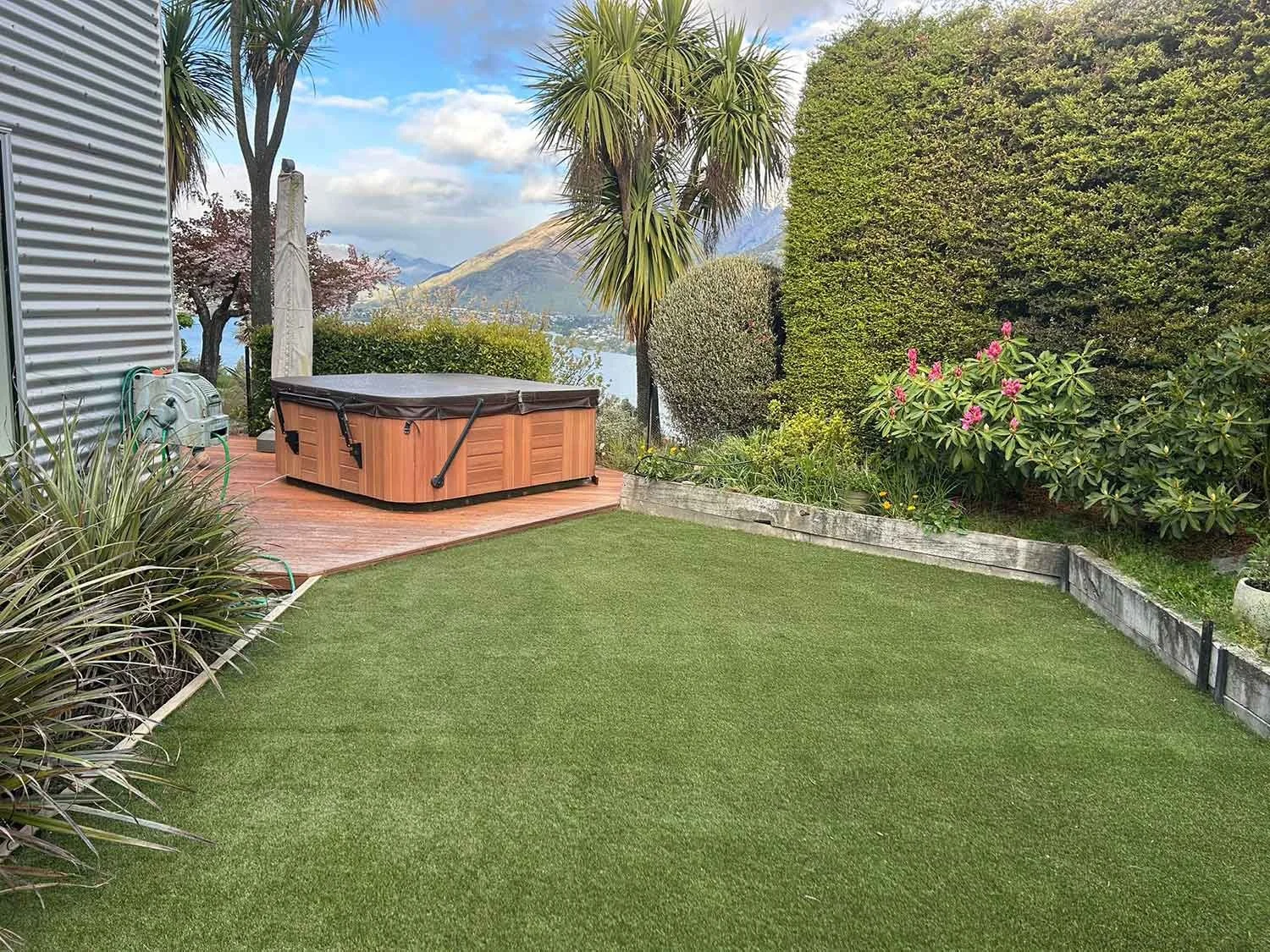 Lush backyard with artificial grass, wooden deck, hot tub, and surrounded by trees and shrubs, with mountains and sky in the background.