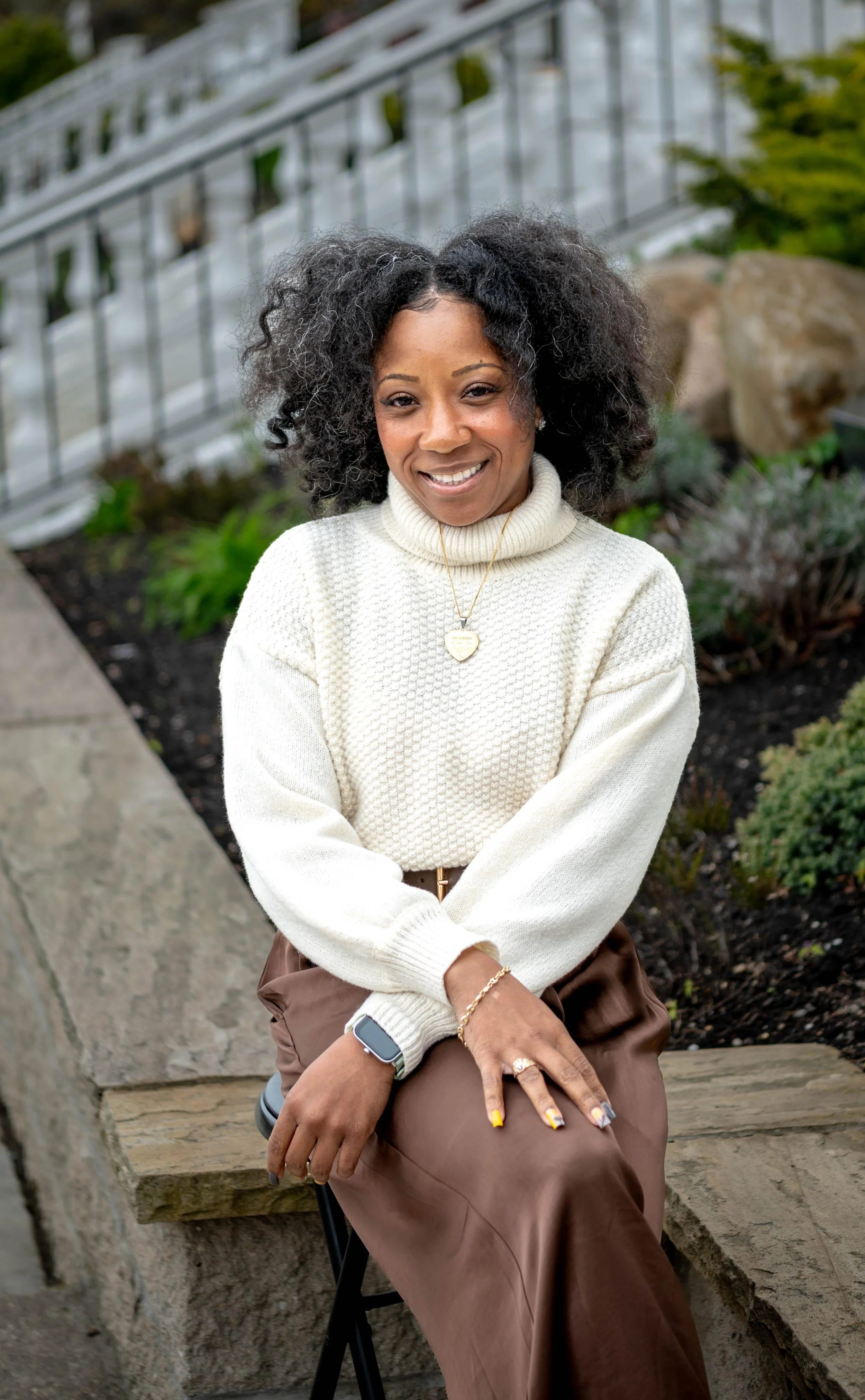 A woman sitting outdoors on a stone bench, wearing a white turtleneck sweater, brown pants, and gold jewelry, with curly dark hair and a smile, in front of a garden with rocks and plants.