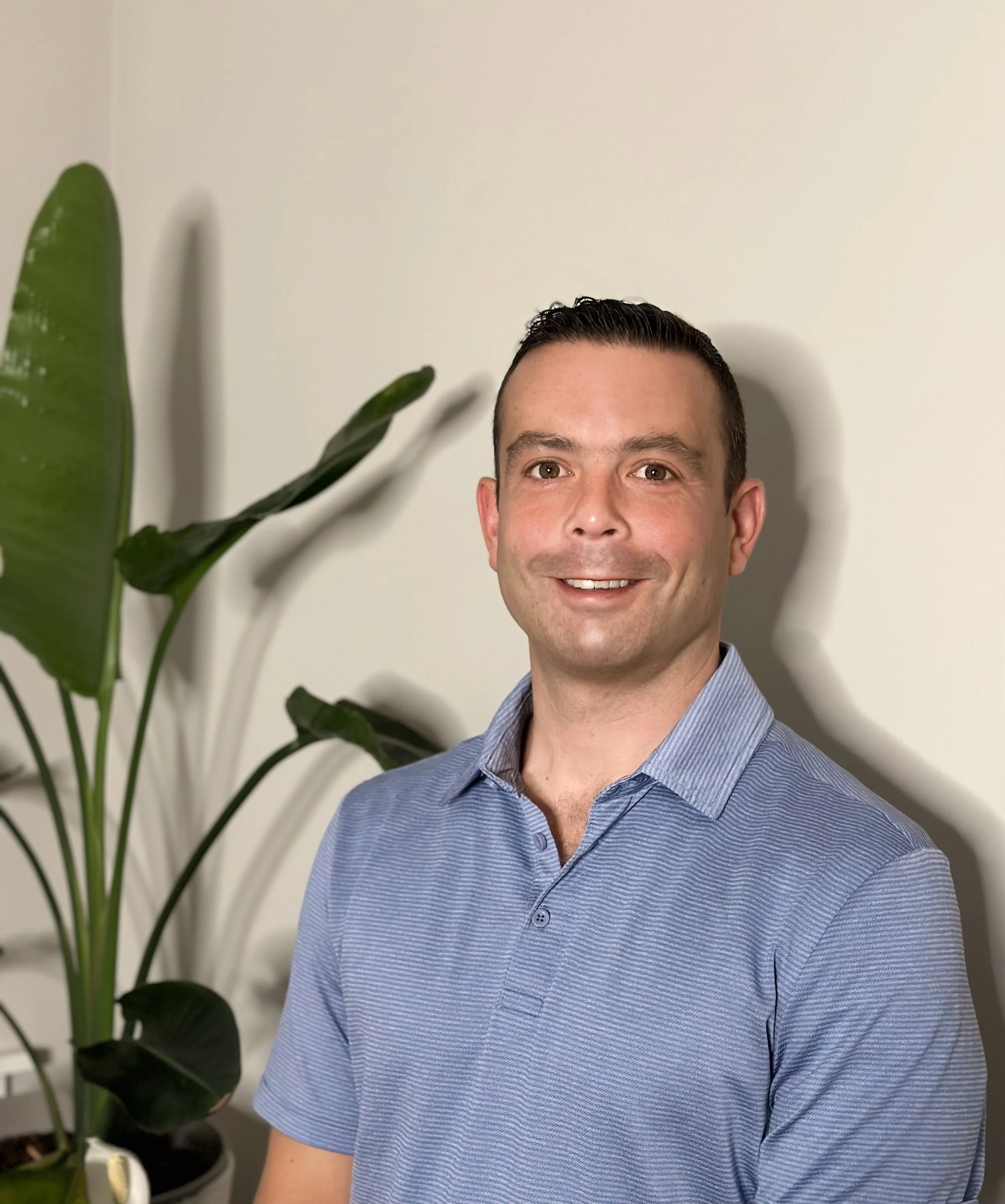 A smiling man with short dark hair, wearing a blue striped button-up shirt, in front of a white wall with a large green plant next to him.