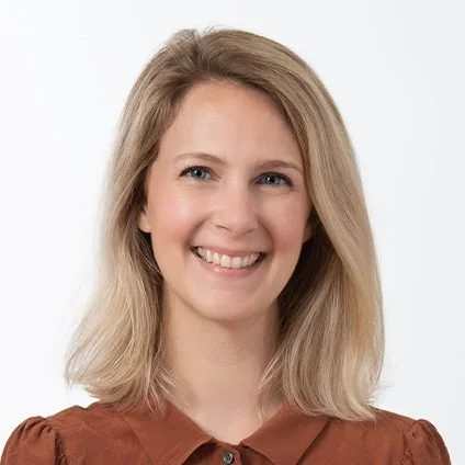 Portrait of a smiling woman with shoulder-length blonde hair, wearing a brown top, against a white background.