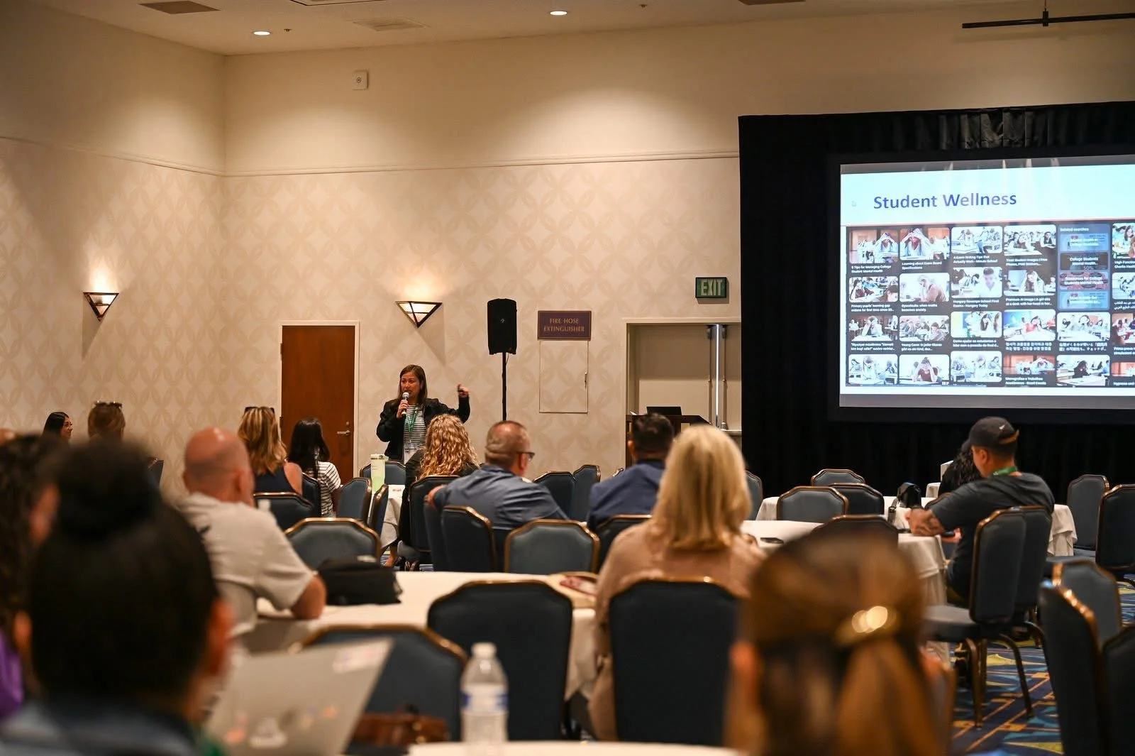 Aileen Dinkjian giving a presentation at a conference, standing in front of a large screen that displays the title 'Student Wellness' with various images and text. The audience is seated at round tables in a conference room.