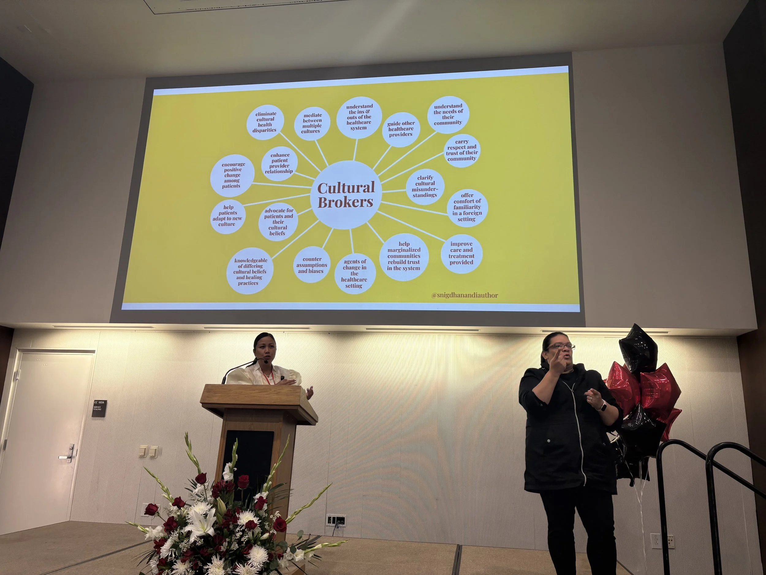 A woman stands at a wooden podium, speaking into a microphone, in front of a large screen displaying a diagram titled 'Cultural Brokers'. Another person stands to the right of the podium, holding a bunch of balloons, and appears to be signing or speaking. There is a floral arrangement at the base of the podium.