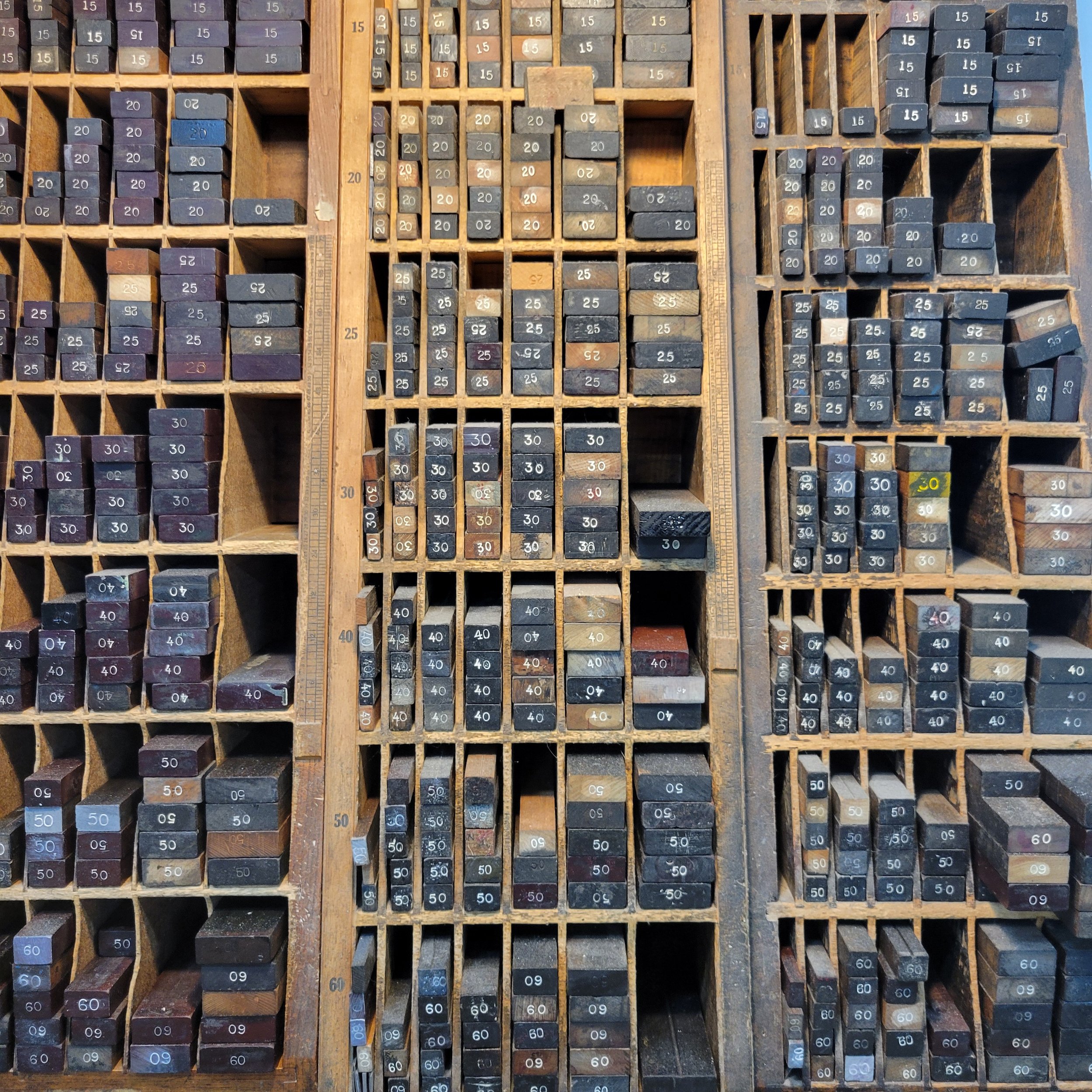 A wooden drawer with multiple compartments containing rubber stamps in various sizes, with numbers like 15, 20, 25, 30, 40, 50, and 60 printed on top. The stamps are arranged in rows and columns, varying in color from black, purple, blue, to brown.