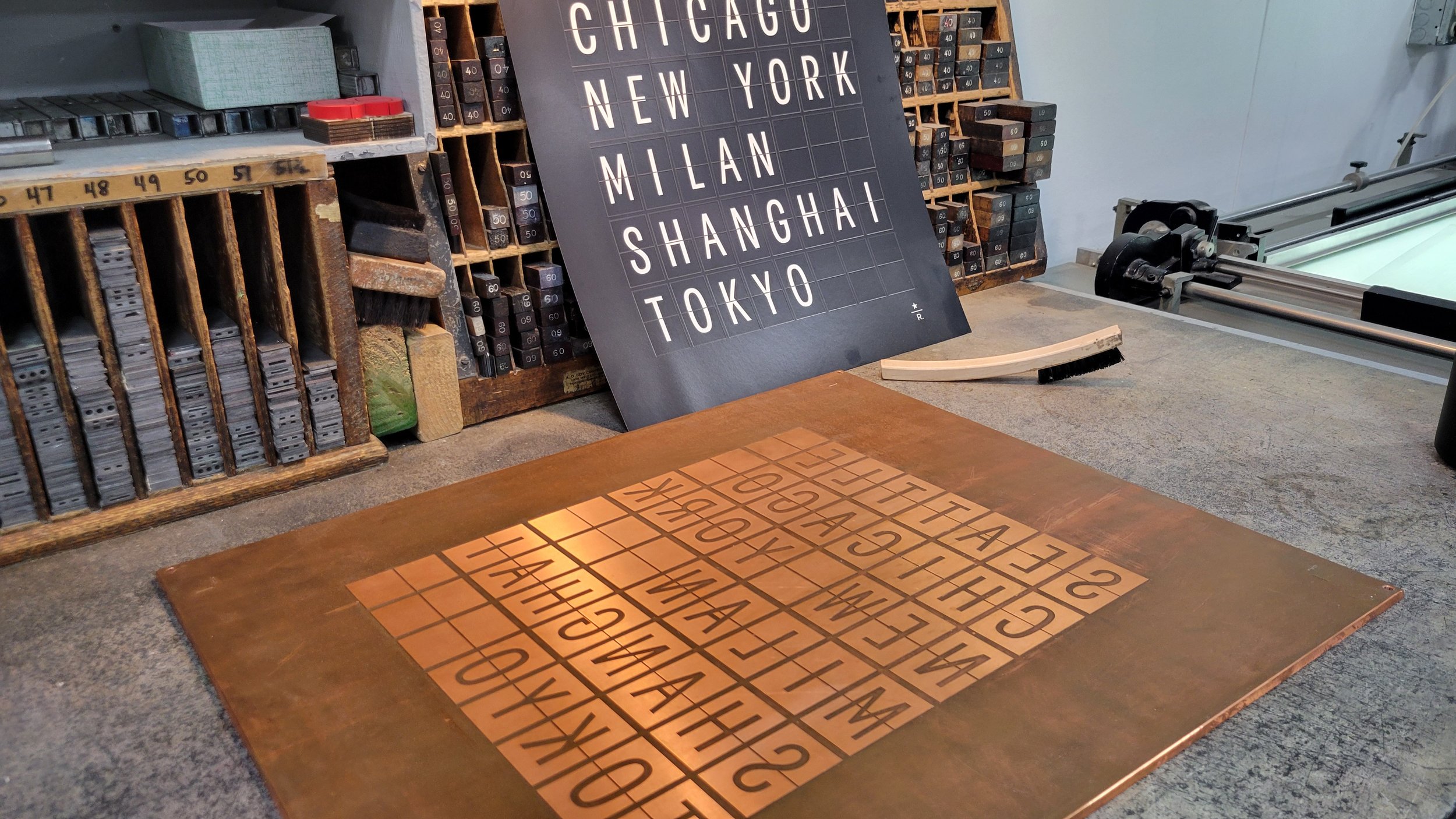 A worktable with metal letter stamps and a rubber mat with alphabet letters. A sign with city names is in the background, and shelves with more letter stamps are visible.