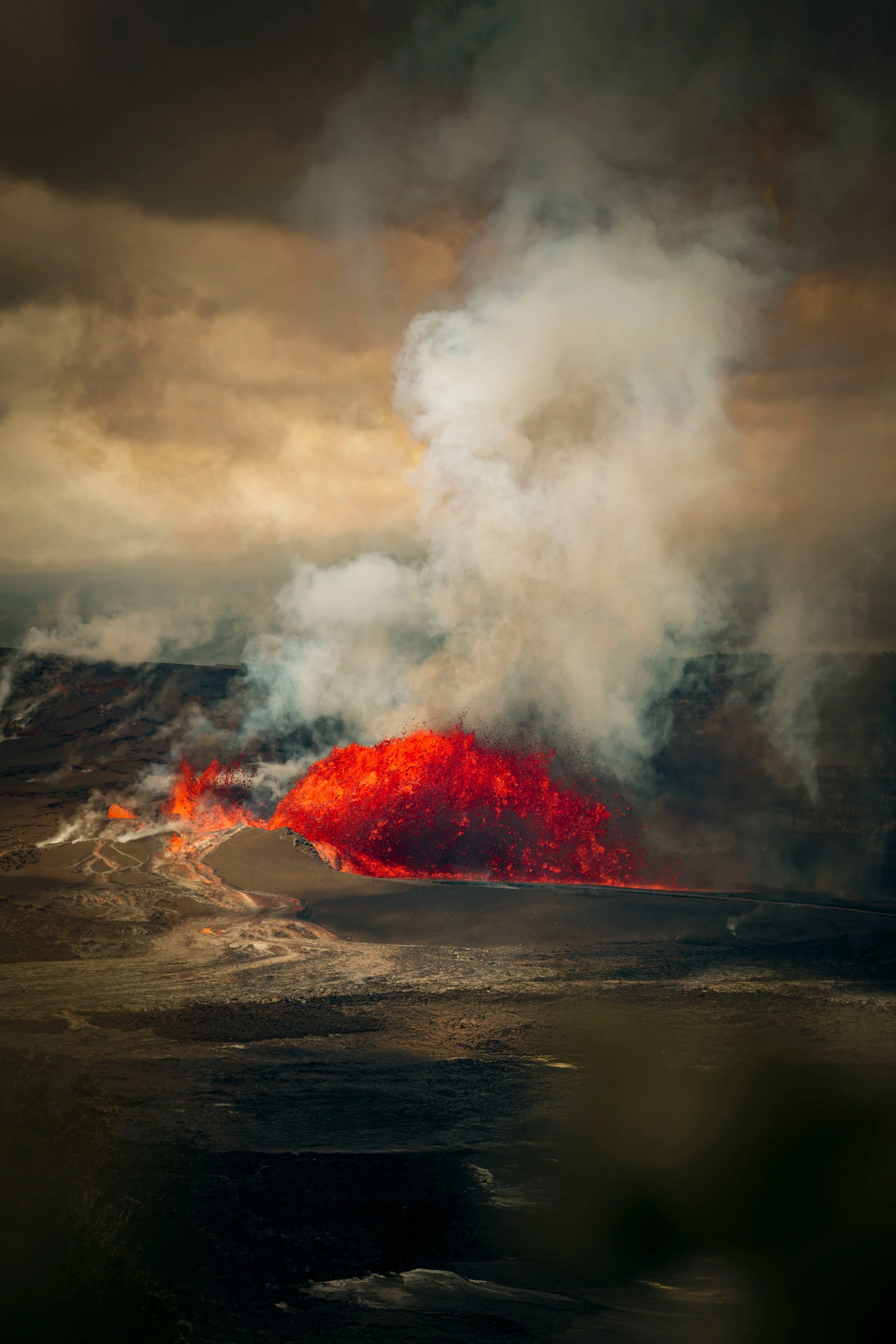 Red lava splashing out of a brown gray mountainous landscape, with smoke rising into a gray clouded sky. Photo by Jonathan Ikemura via Unsplash