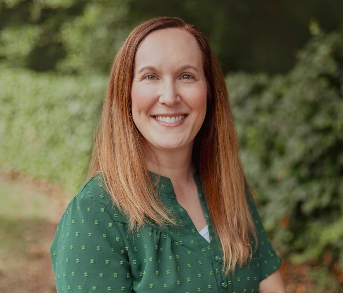 A woman with long reddish-brown hair smiling outdoors, wearing a green blouse with small yellow patterns, surrounded by greenery.