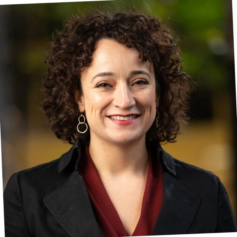 woman with dark curly hair, black jacket, wine colored blouse