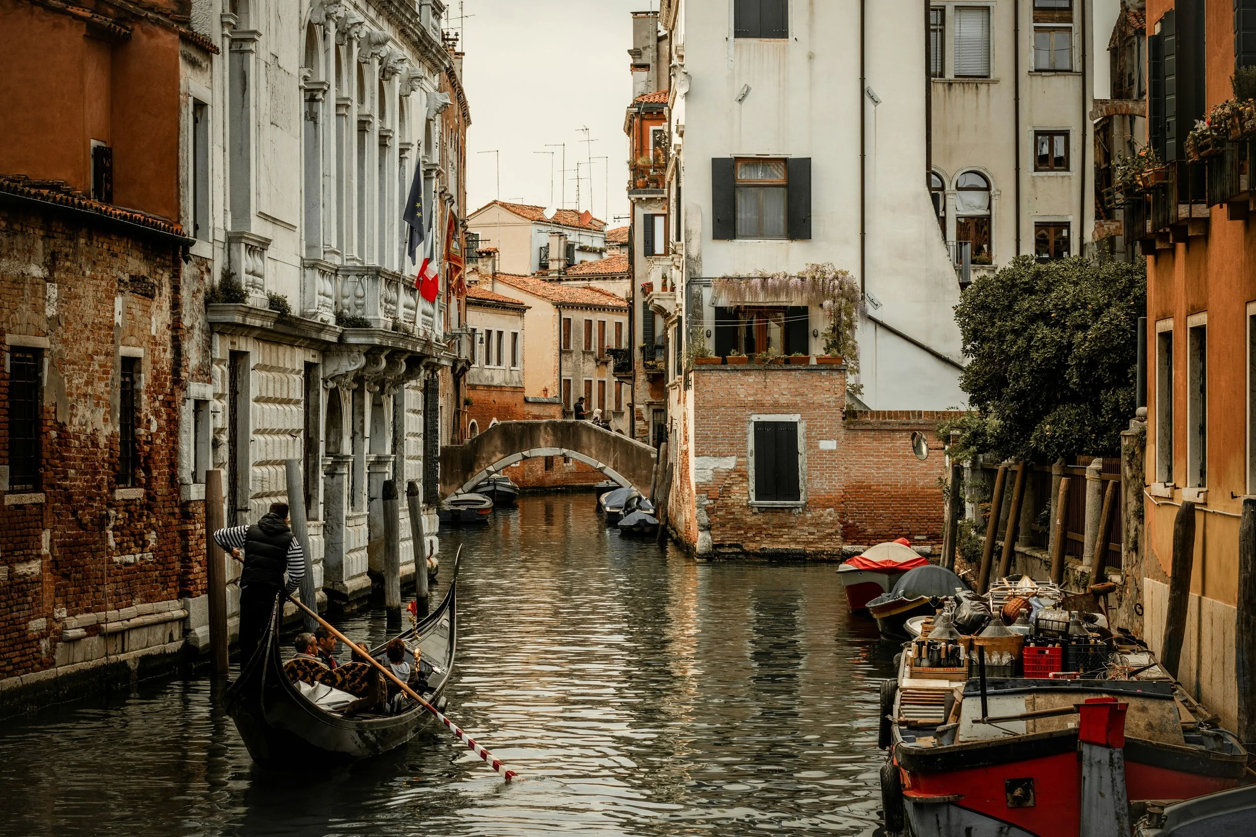 Gondola navigating a narrow canal in Venice, Italy, with historic buildings on either side, some with balconies and shutters, and small boats docked along the canal.