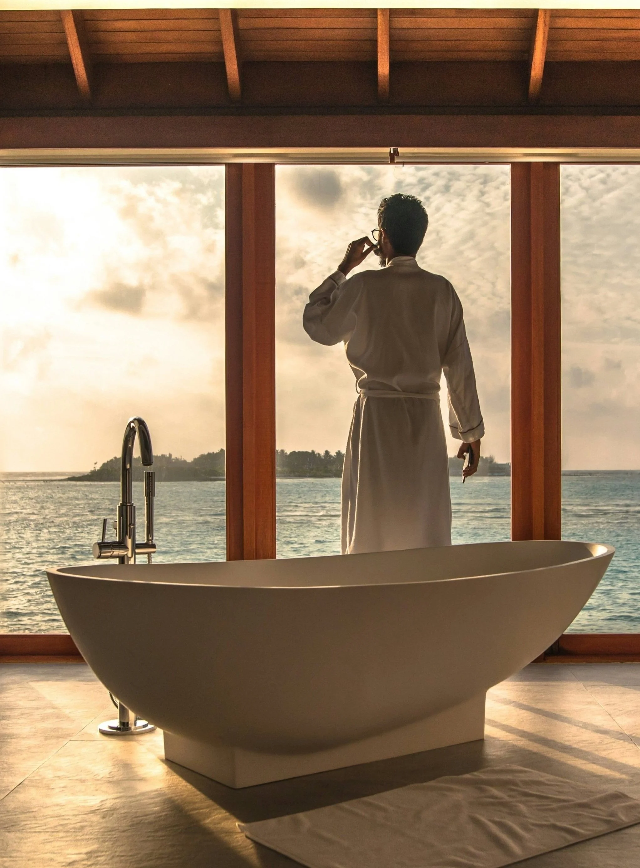 Man in bathrobe standing by large window overlooking ocean at sunset, with freestanding bathtub in foreground, and wooden ceiling.