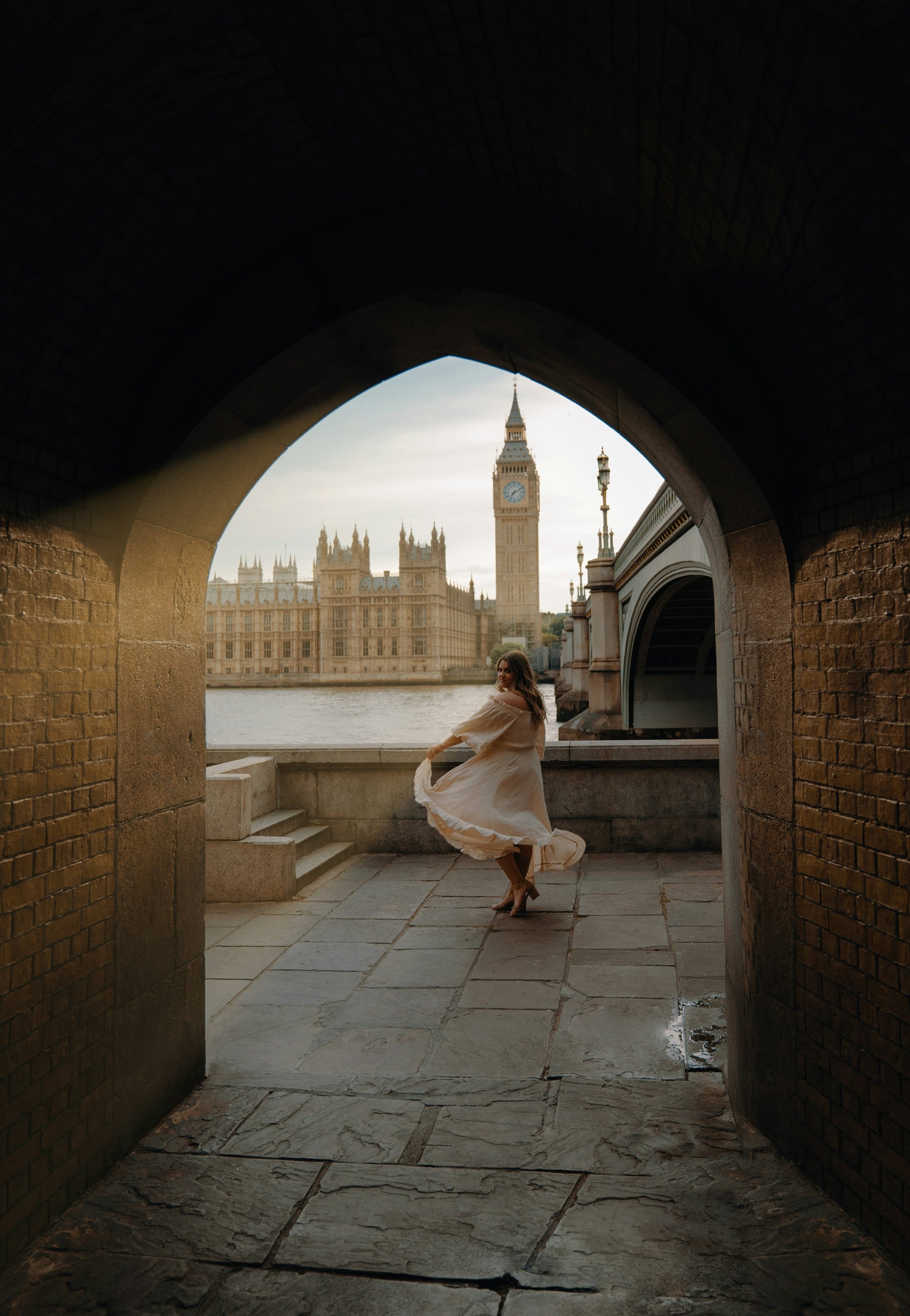 A woman in a flowing beige dress spinning and dancing in a tunnel with brick walls, overlooking the River Thames, the Palace of Westminster, and Big Ben in the background.