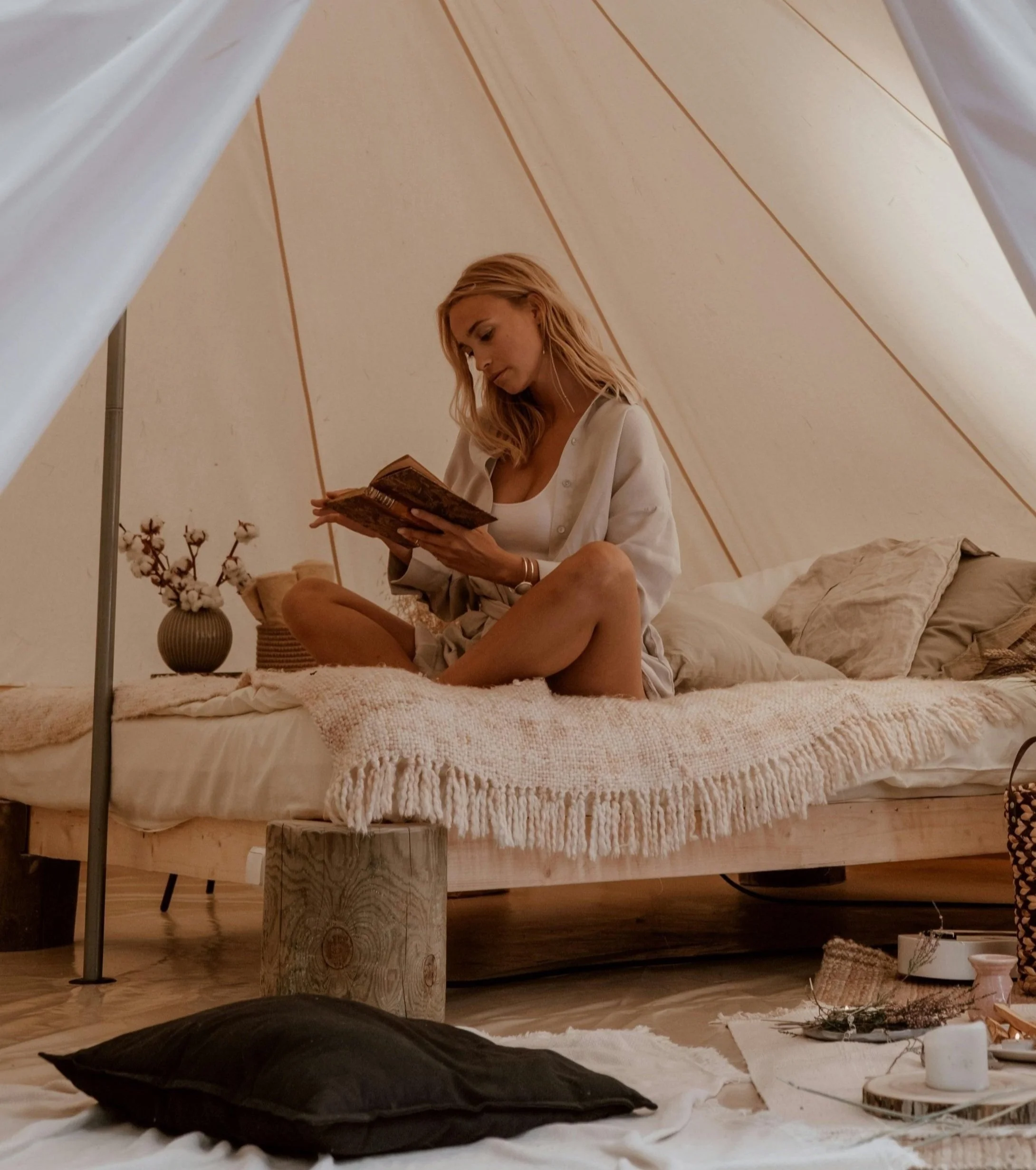 A woman with blonde hair sitting on a bed in a cozy, softly lit room, reading a book. The room has neutral tones, with a wooden bed frame, pillows, a blanket, and decorative items including a vase with cotton branches and candles.