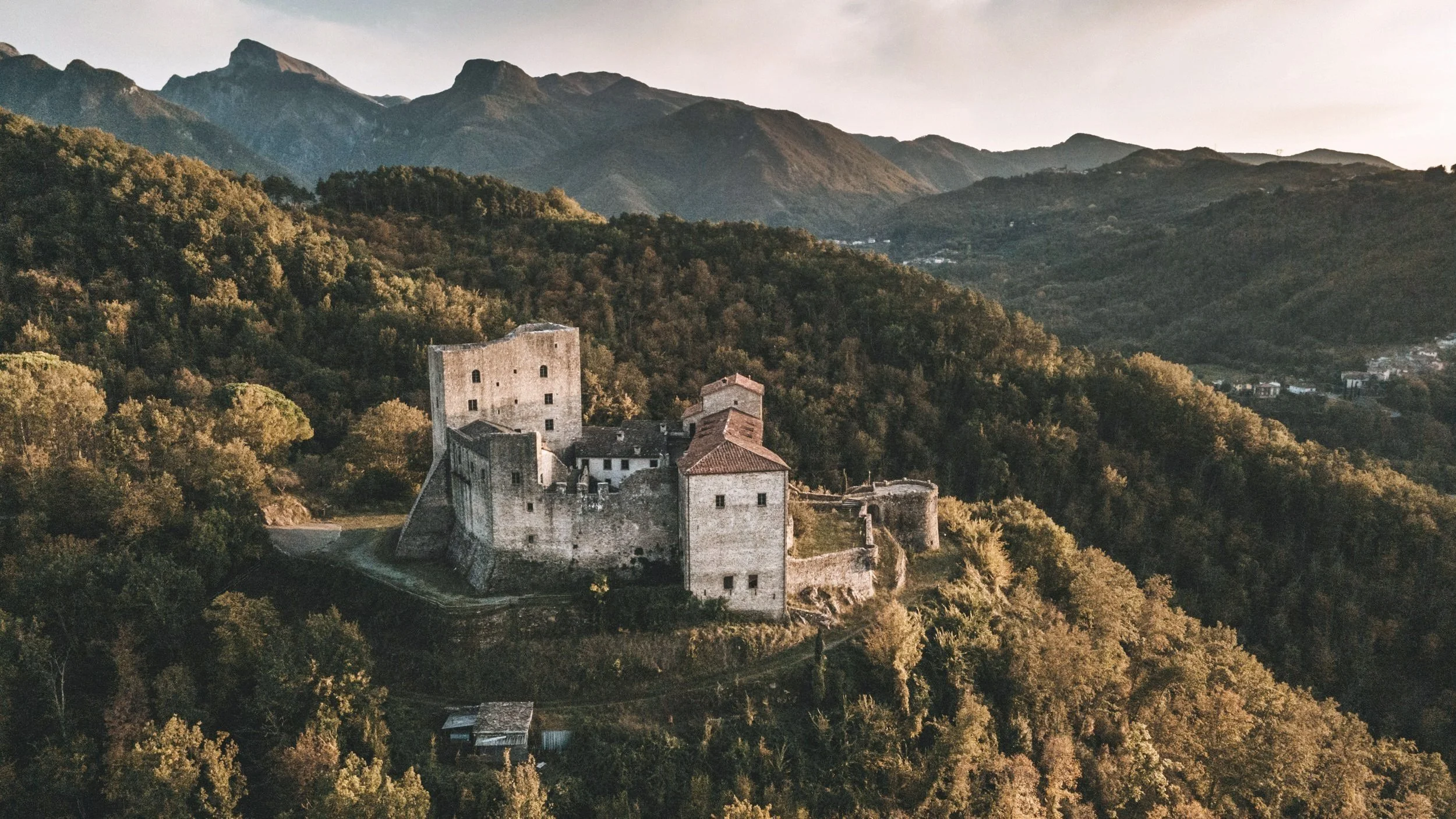 A historic stone castle on a hill surrounded by dense green forest, with mountains in the background under a cloudy sky.