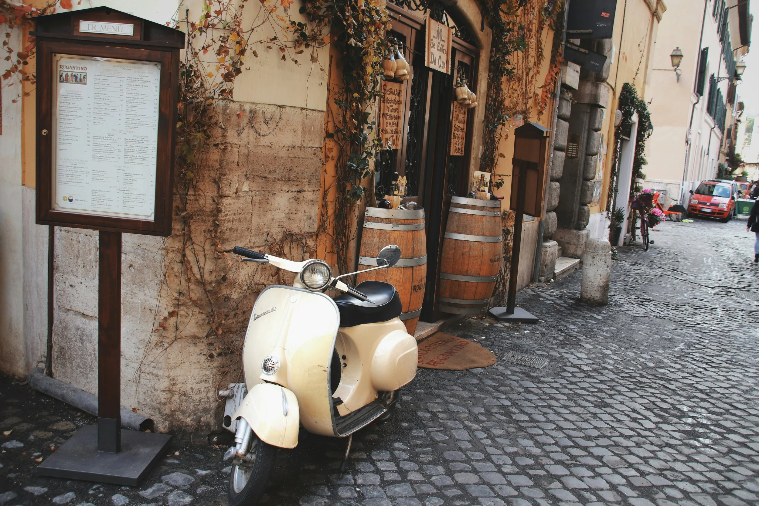 A narrow street scene with a vintage cream-colored scooter parked outside a restaurant. The restaurant has a menu displayed on a wooden frame, and some hanging decorations are visible beside its door. The street is paved with cobblestones and shows a parked red car further down the street. Buildings along the street have shutters and potted plants, with vines climbing the walls.