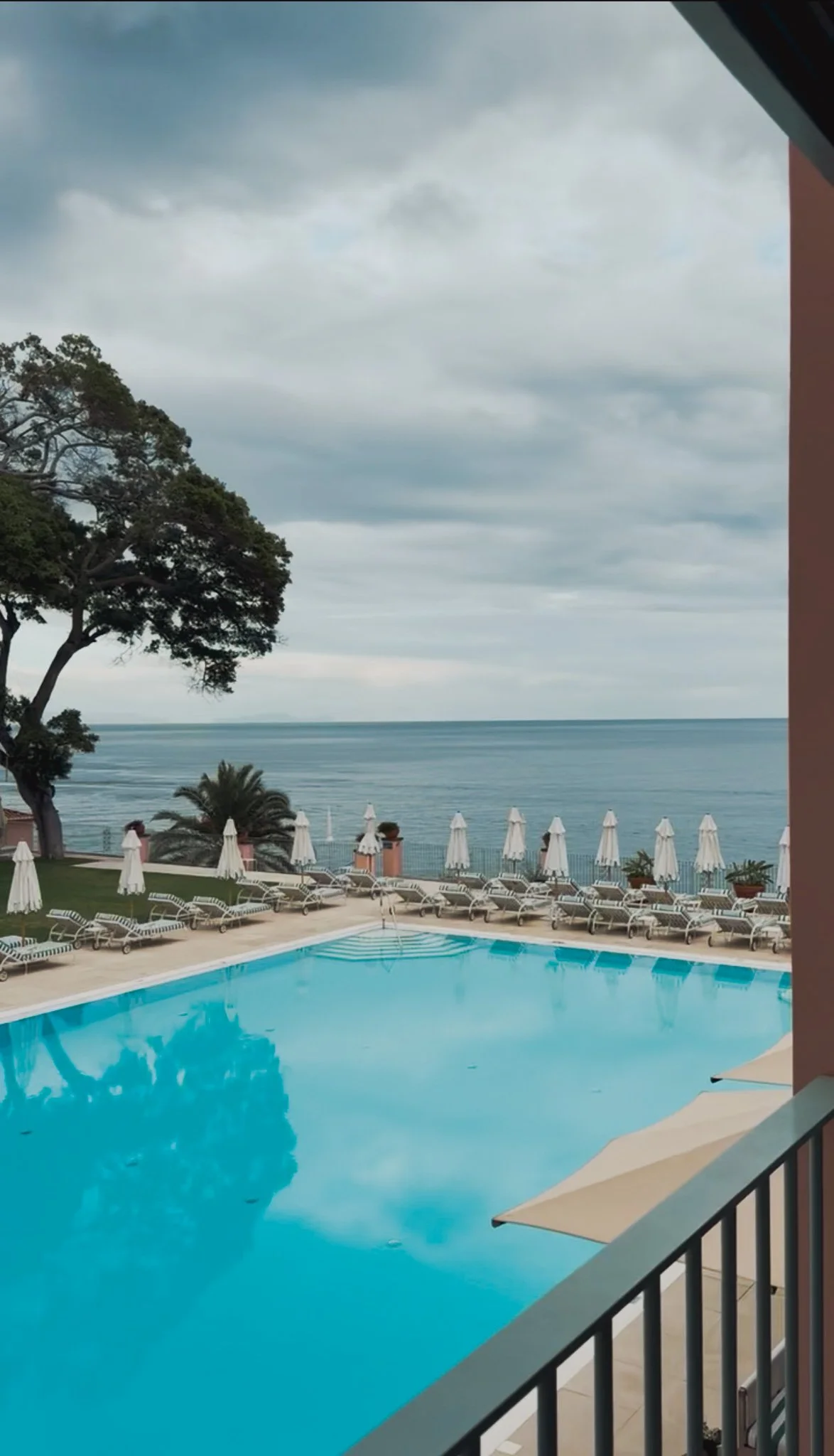 A view of a swimming pool with lounge chairs and umbrellas, overlooking the ocean and cloudy sky.