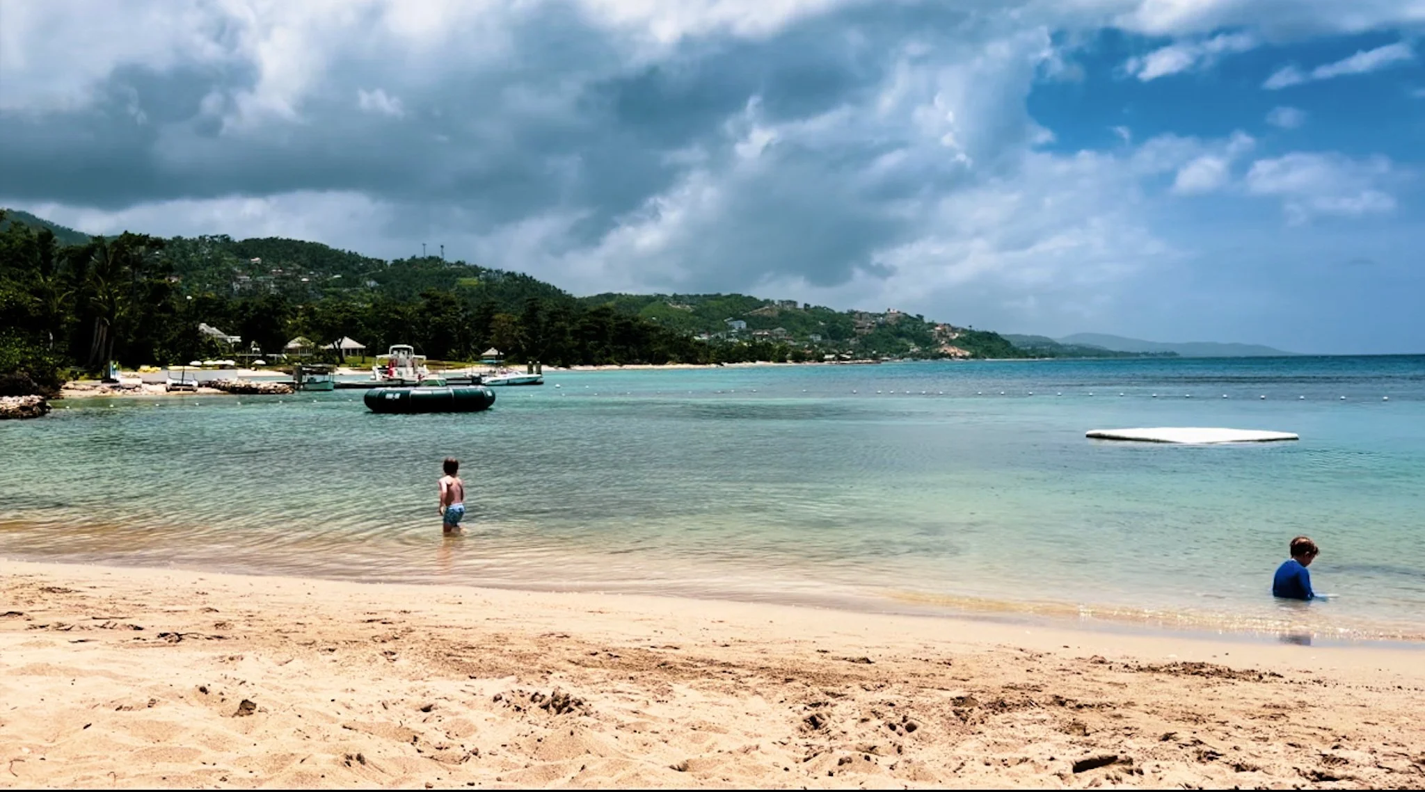 Children playing in shallow ocean water near a sandy beach, with boats docked near the shore and a hillside with houses and greenery in the background under cloudy skies.