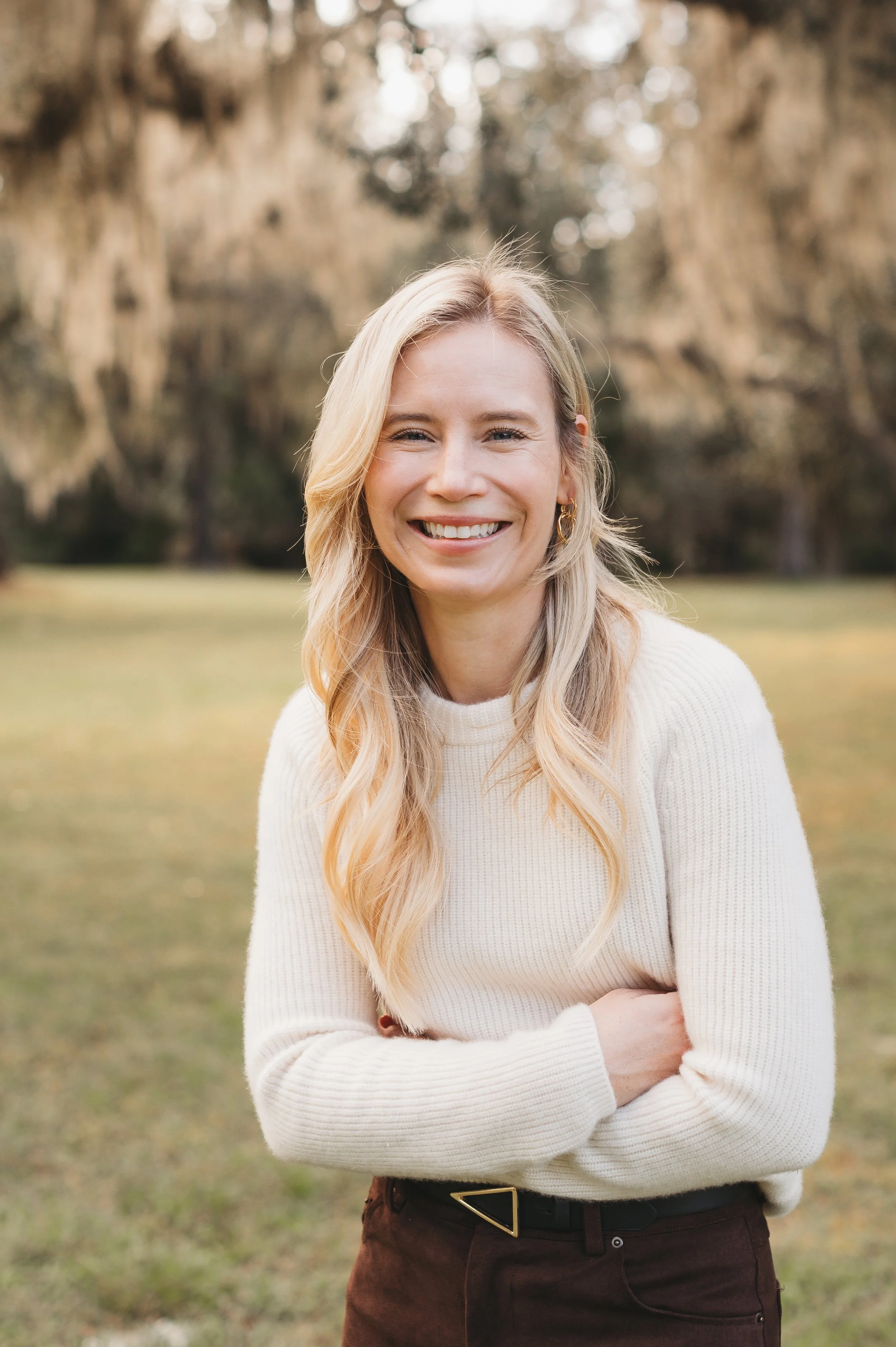 A woman with long blonde hair smiling outdoors with trees in the background, wearing a white sweater and dark pants.