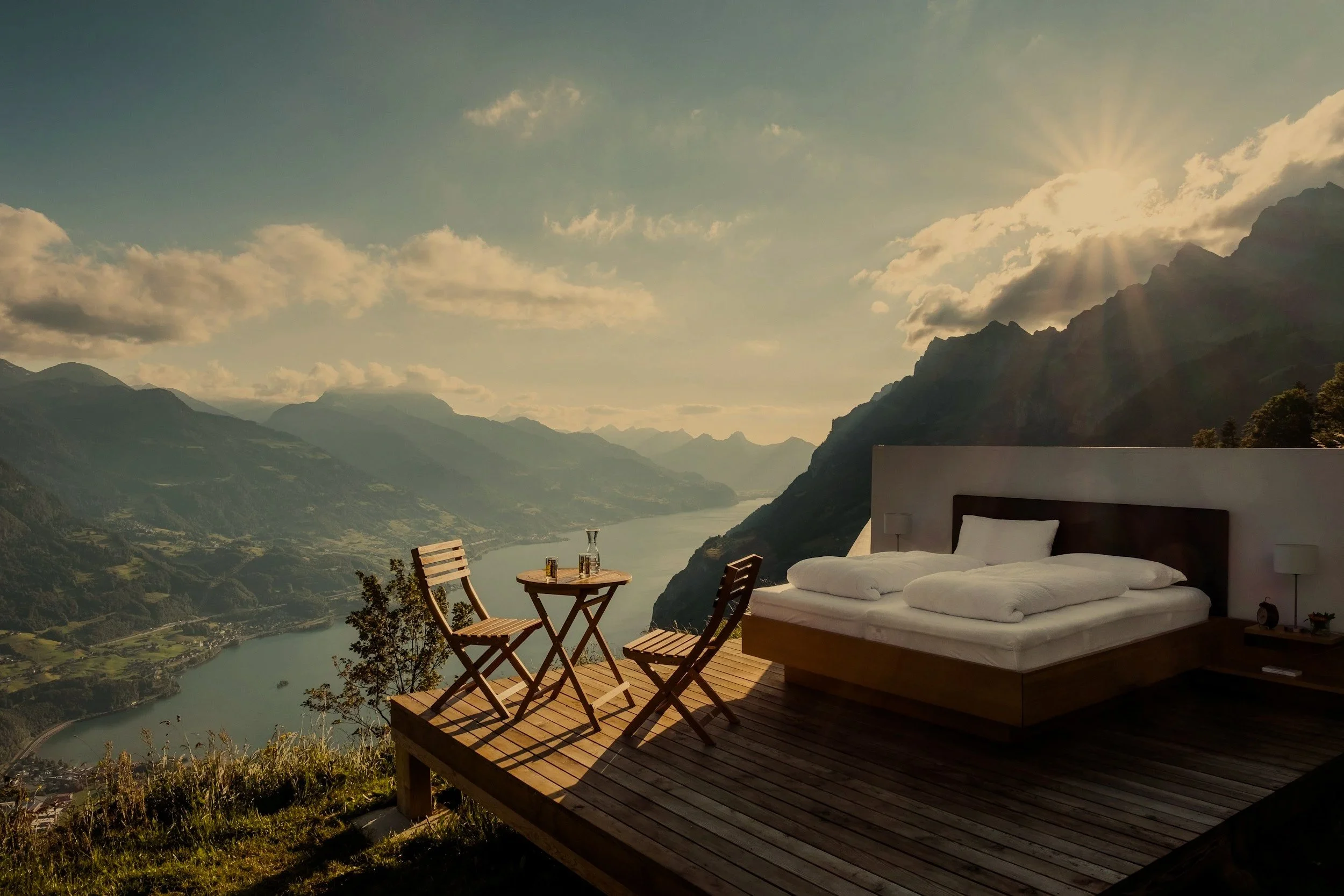 A scenic outdoor deck with a bed, chairs, and a table overlooking a river and mountain landscape during sunset with sun rays and clouds.