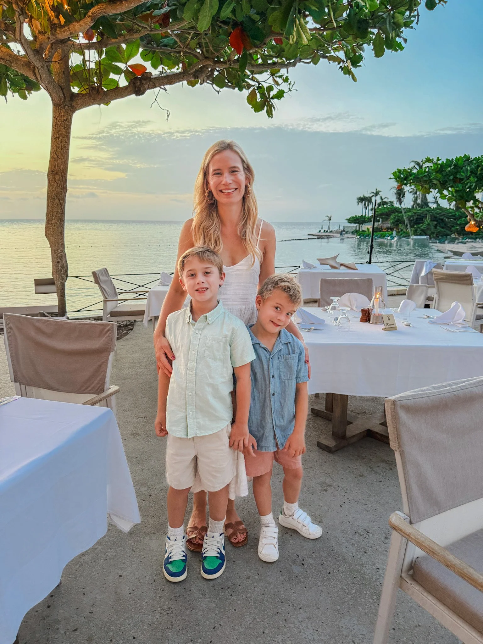 A woman and three young boys standing together at an outdoor restaurant near water, with tables and chairs, under a tree, during sunset or early evening.