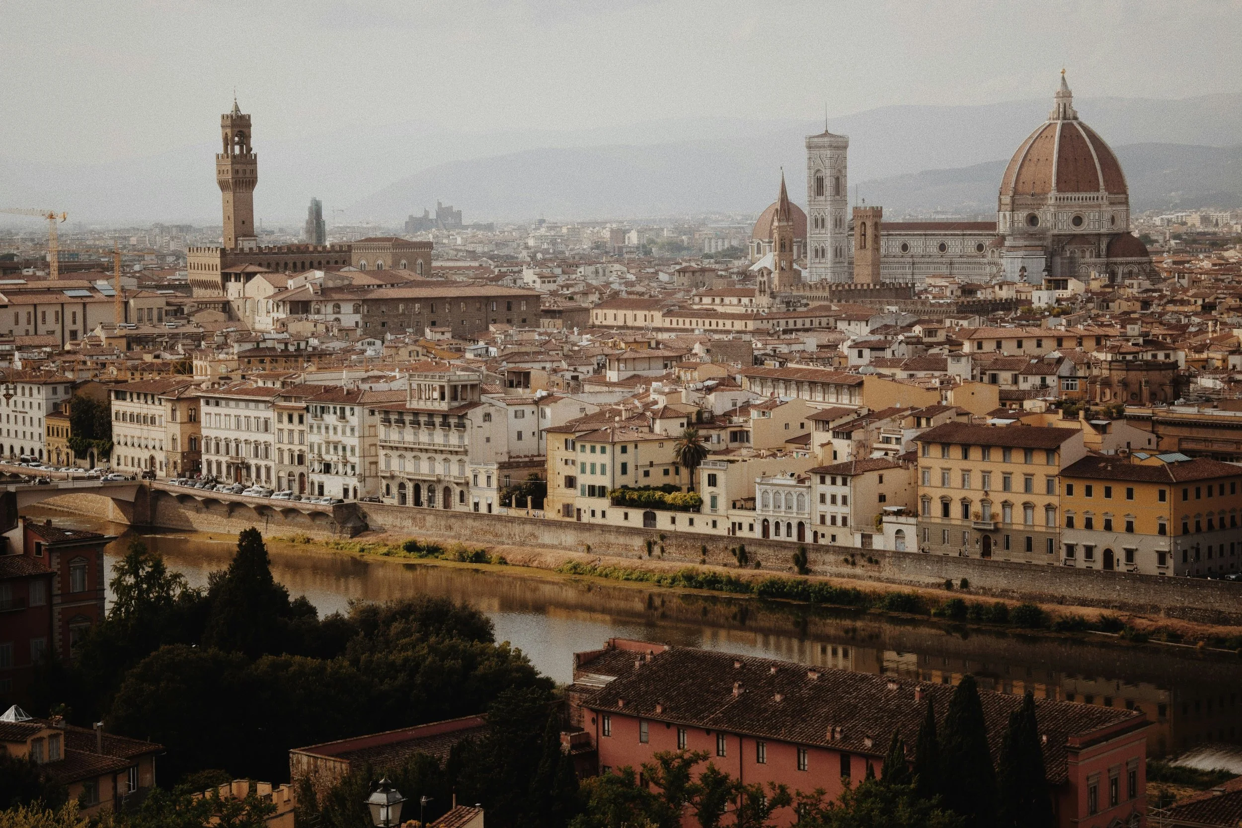 A cityscape view of Florence, Italy including the Florence Cathedral's dome, Giotto's bell tower, and Palazzo Vecchio with surrounding historic buildings and a river in the foreground.