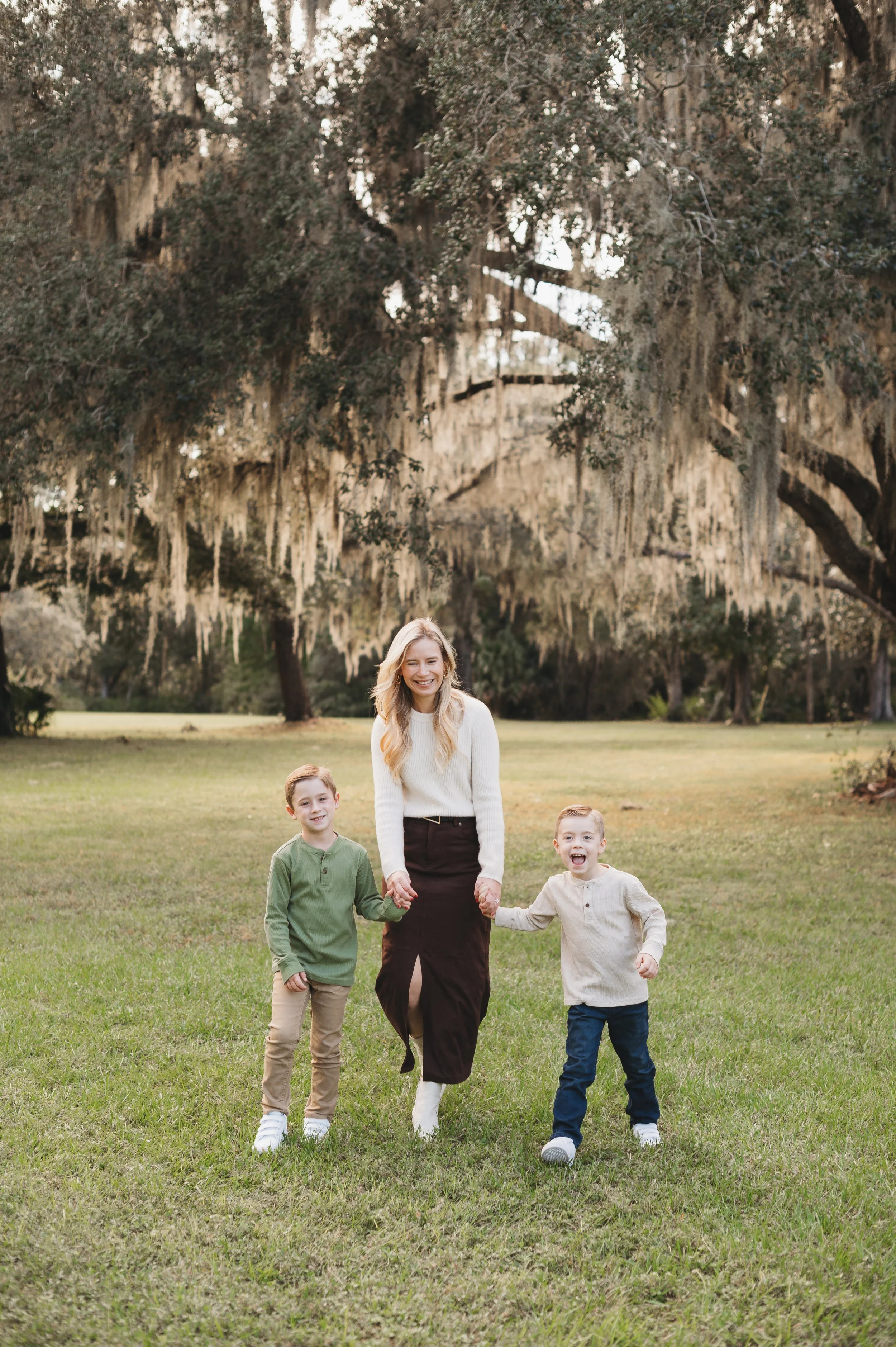 A woman with blonde hair and a white sweater walking hand in hand with three children on a grassy field with large trees in the background.