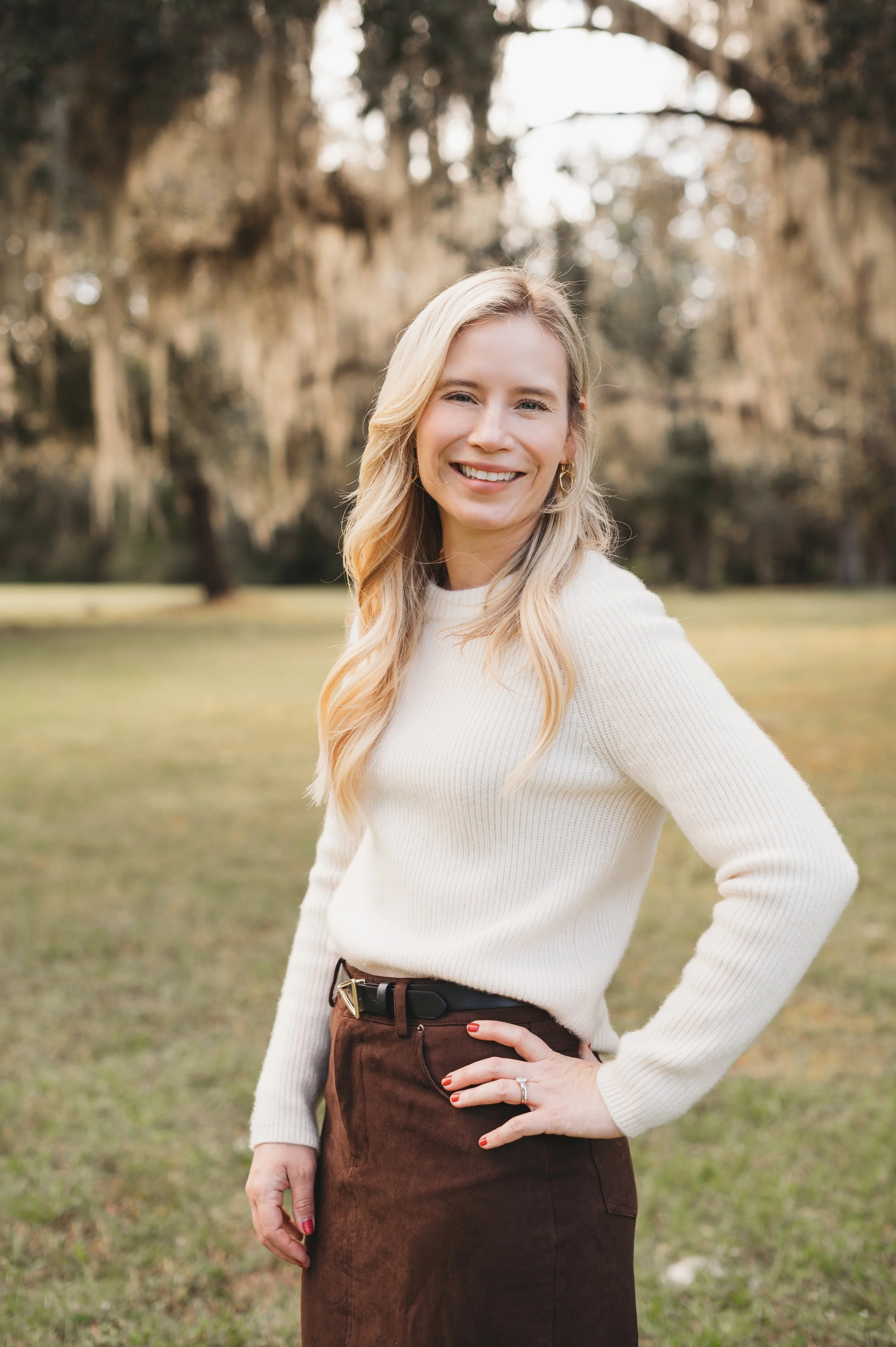 A woman with blonde hair smiling outdoors in a park with trees in the background.