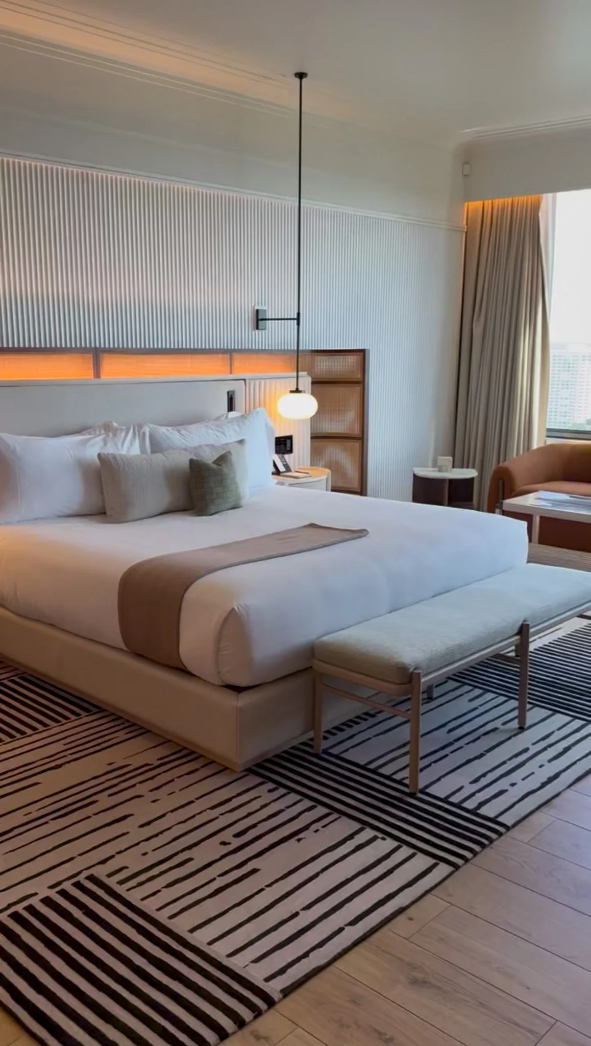 Hotel room with a large bed, side table, bench, and a window with curtains. Modern decor with striped rug and minimalist lighting.