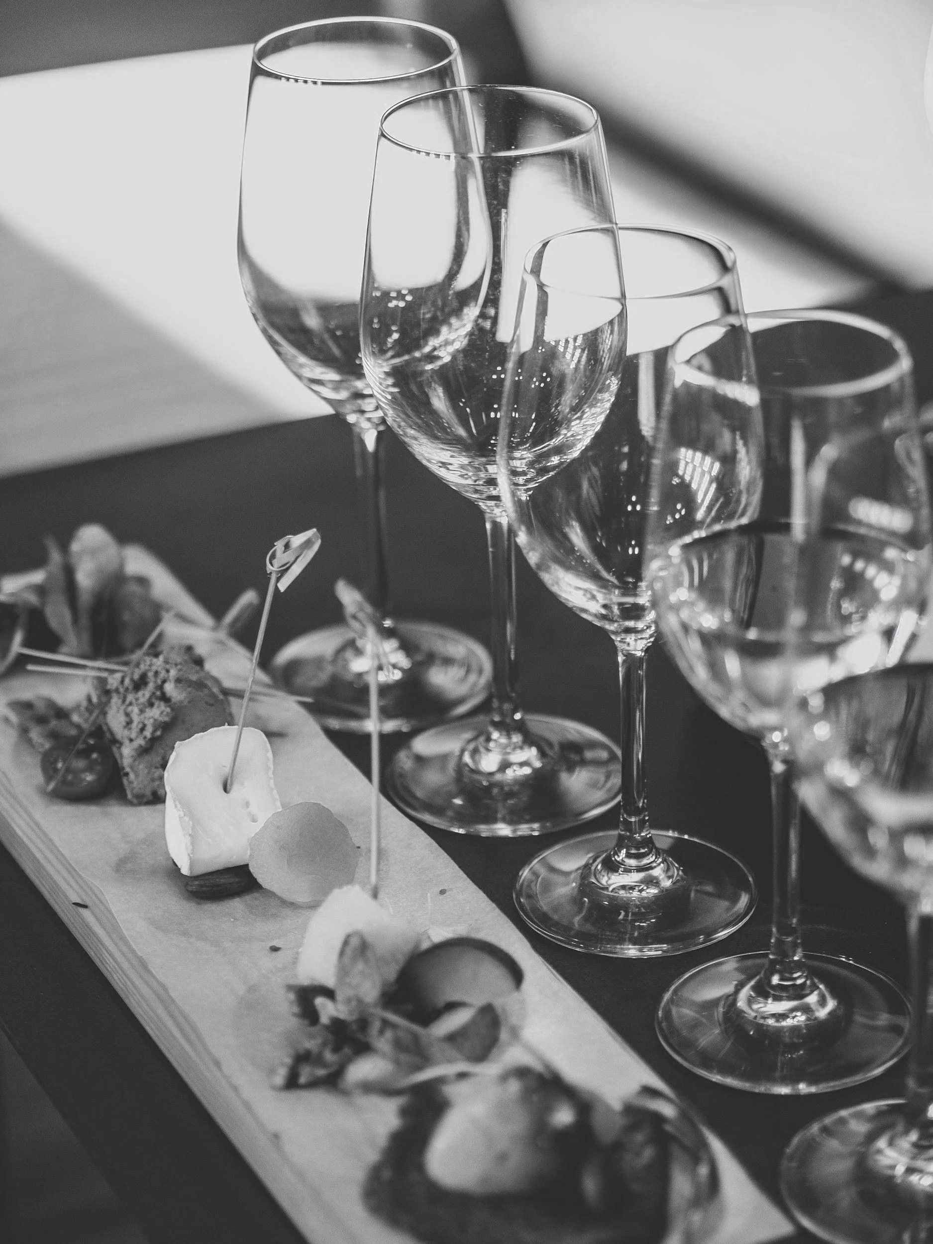 A black and white photo of a flight of wine tasting glasses and a cheese platter with various cheeses, crackers, and fruit.