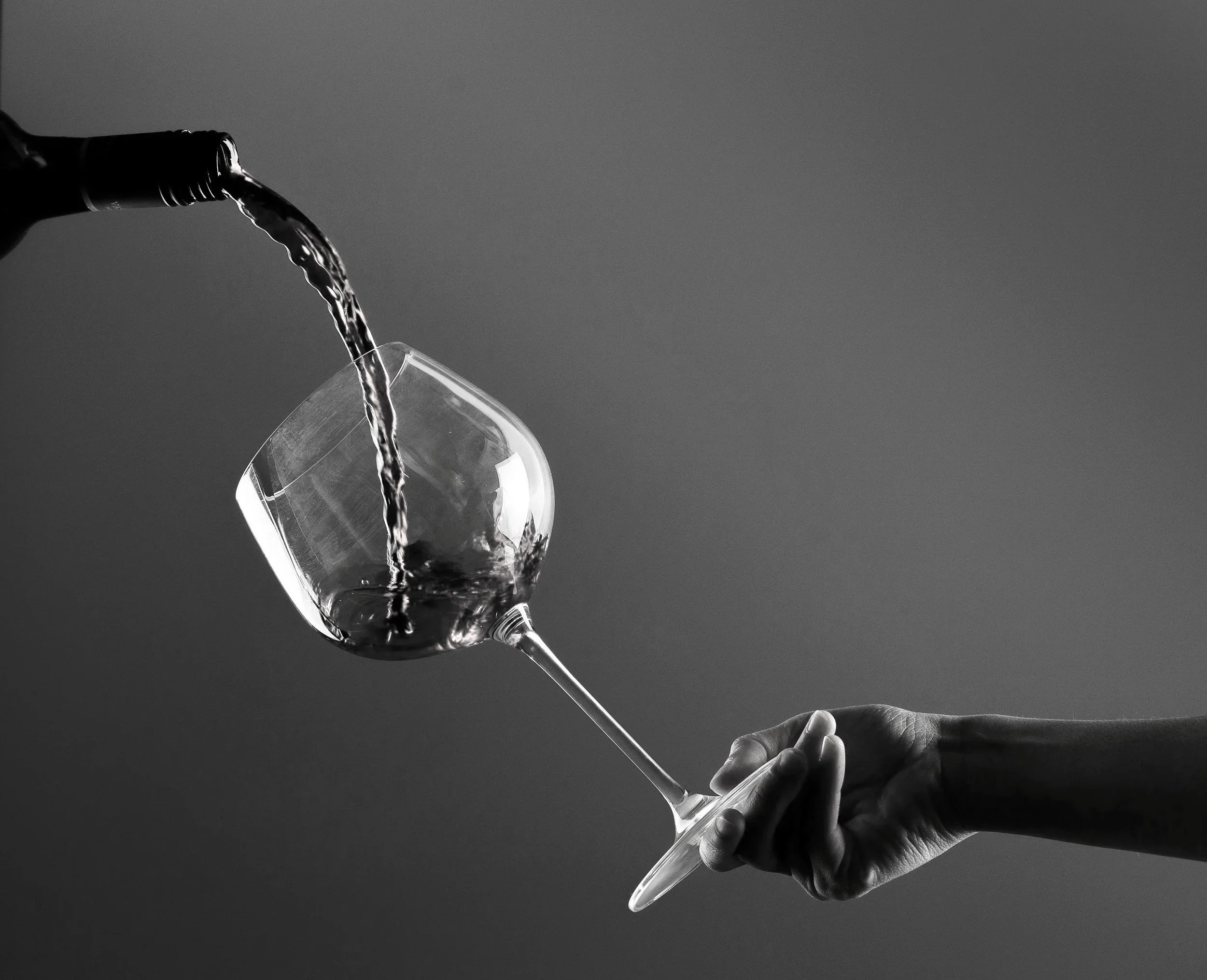 Close-up of a hand holding a wine glass, with another hand pouring wine into it, in black and white.