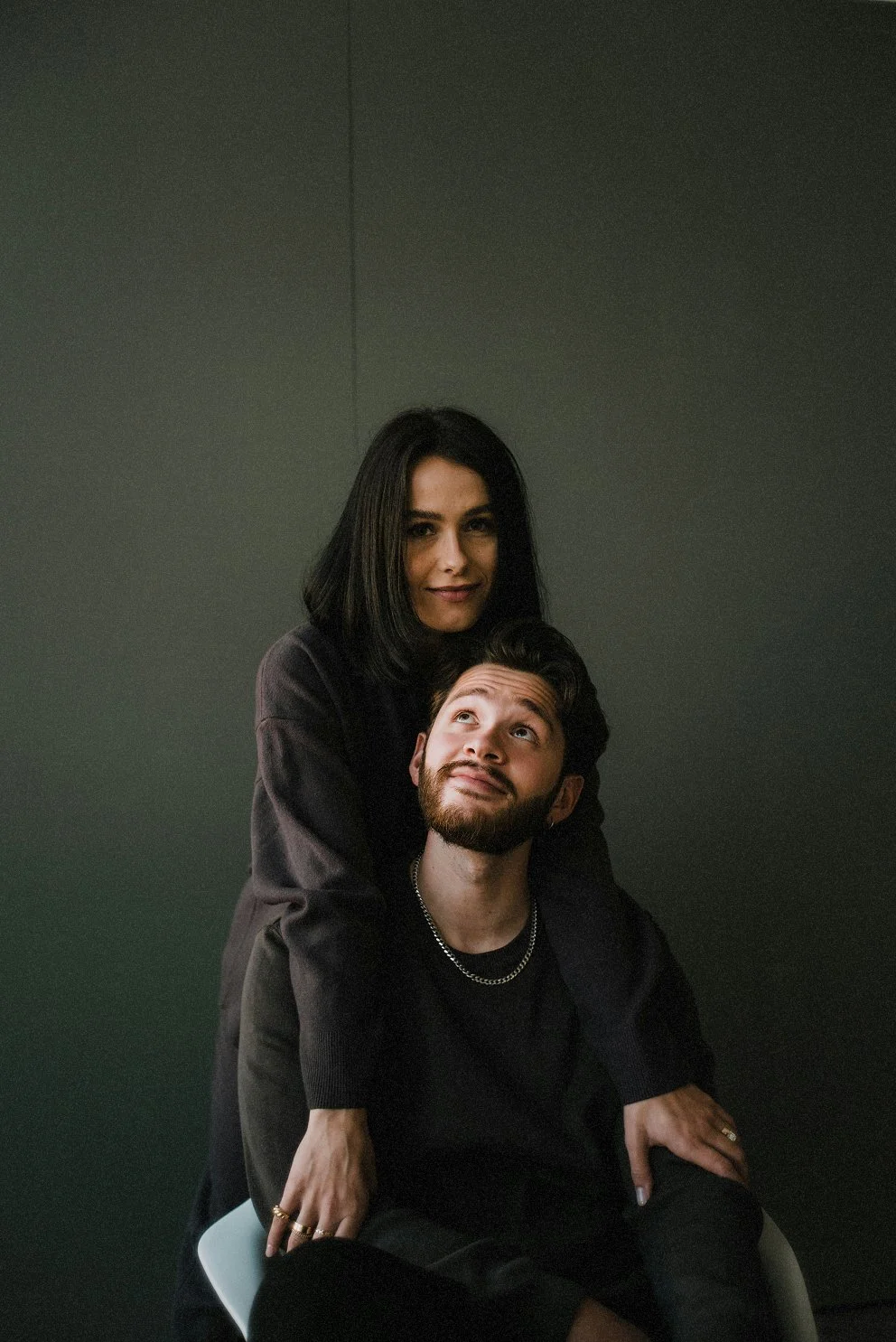 A woman with shoulder-length dark hair embraces a man with dark hair and a beard, both dressed in dark clothing, set against a plain dark background.