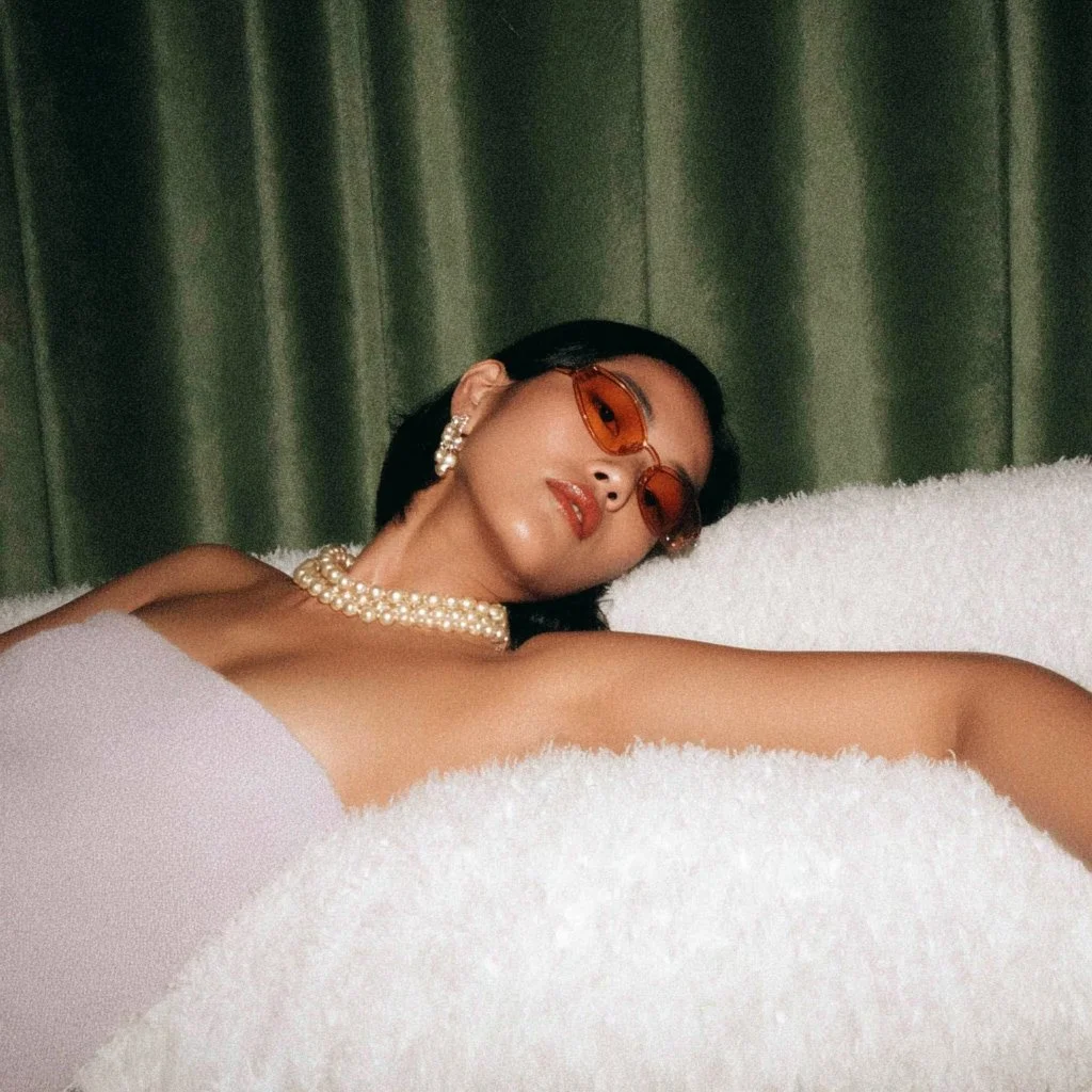 A woman wearing orange-tinted glasses, pearl earrings, and a pearl necklace, lying on a white fluffy surface with a green curtain background.