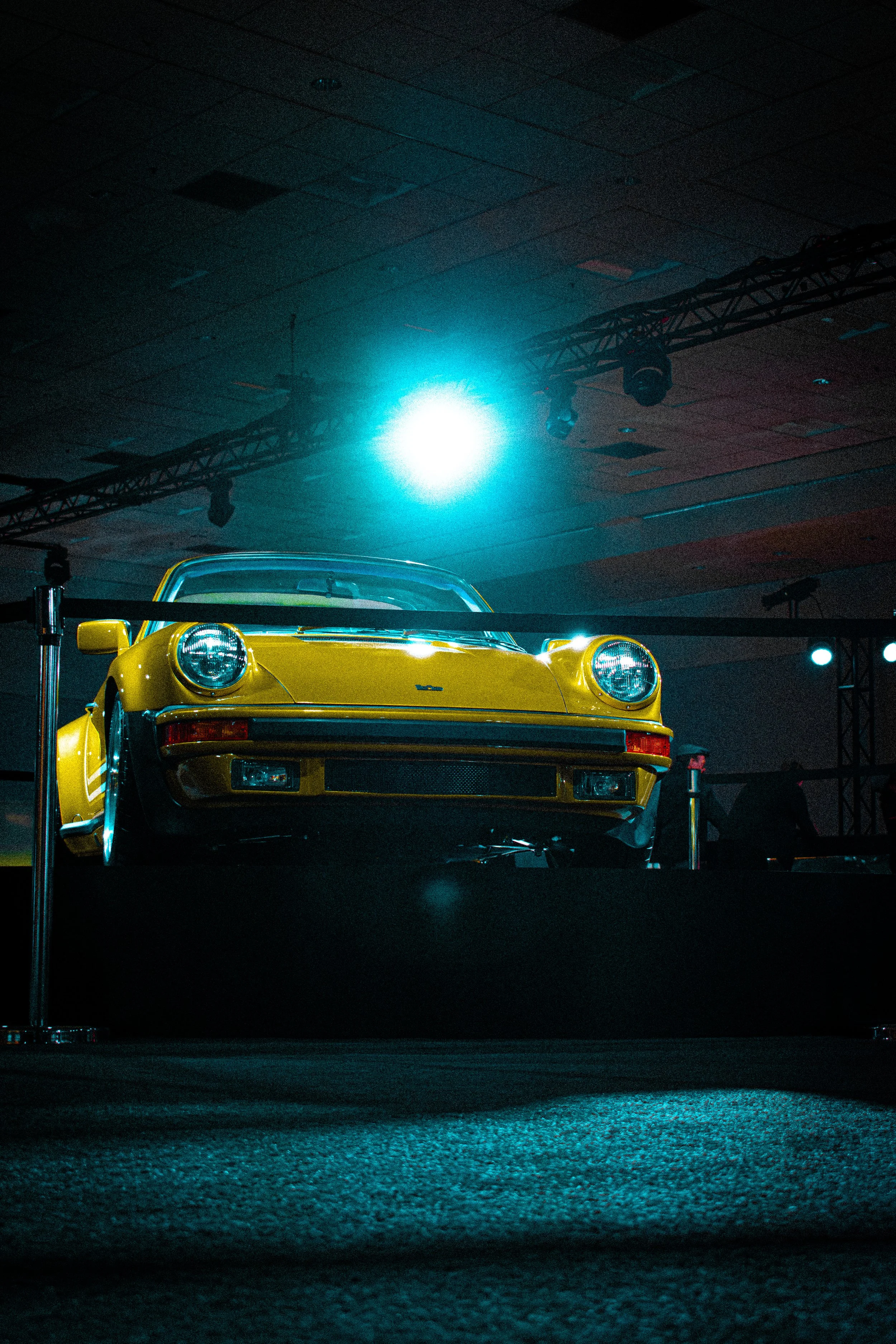 A yellow vintage car displayed on a platform at an indoor auto show, with blue lighting overhead.