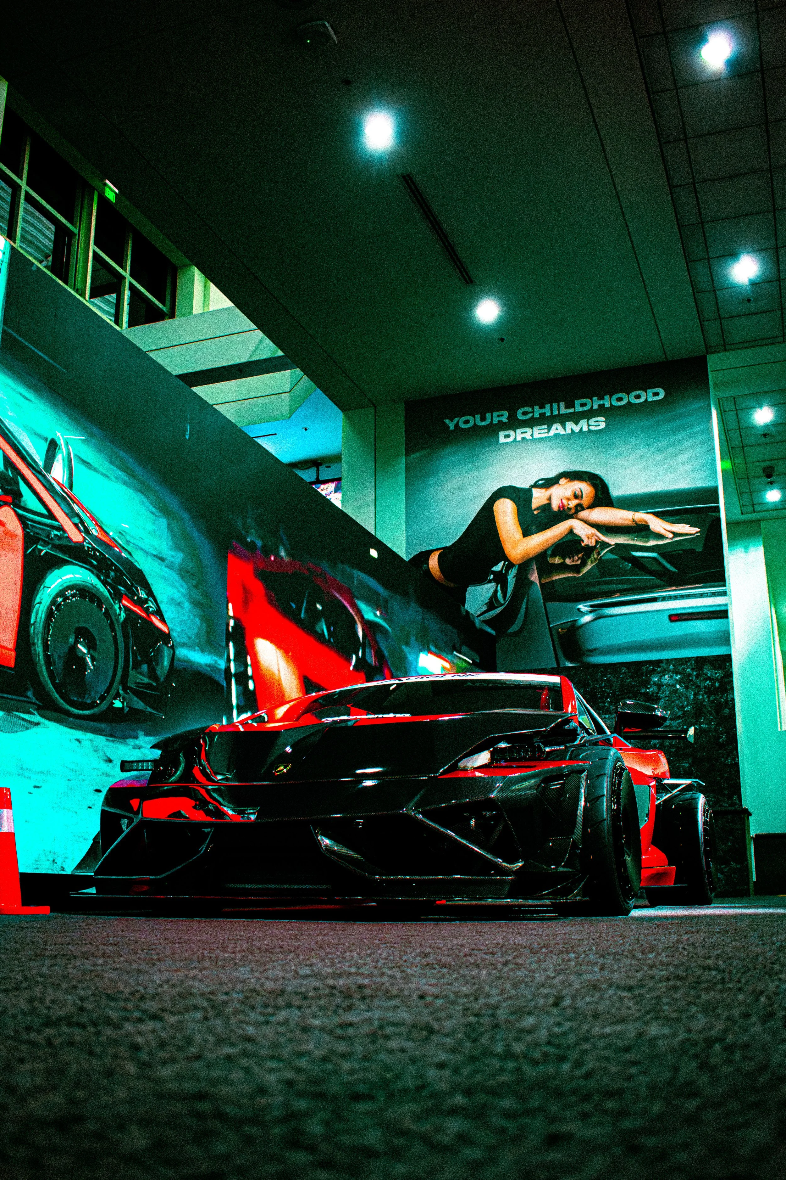 Display of racing car on exhibition floor with promotional posters and a large billboard in the background showing a woman leaning on a car and the words 'Your Childhood Dreams.'