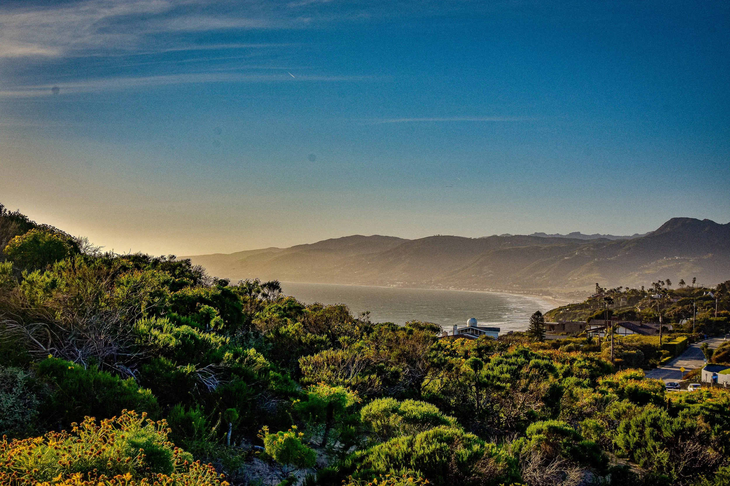 Scenic view of a coastal landscape with lush green bushes in the foreground, a hilltop observatory or lighthouse near the sea, and distant mountains under a clear blue sky with wispy clouds.