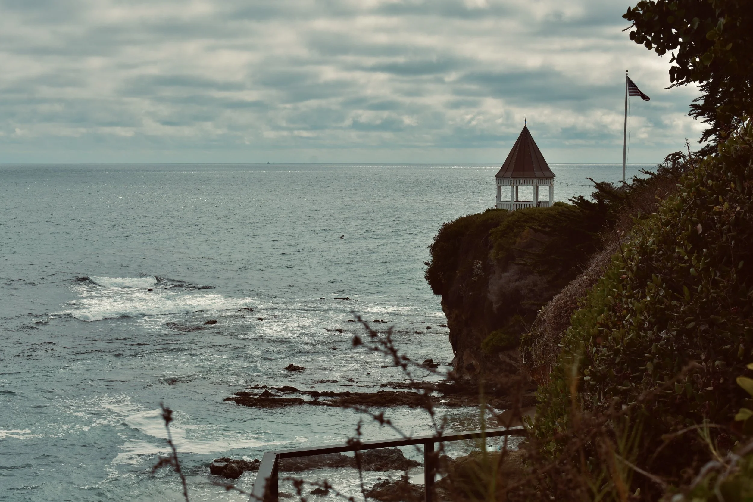 A view of a rocky coast with waves crashing, a small pavilion with a red roof on a cliff, and an American flag flying next to it under cloudy skies.