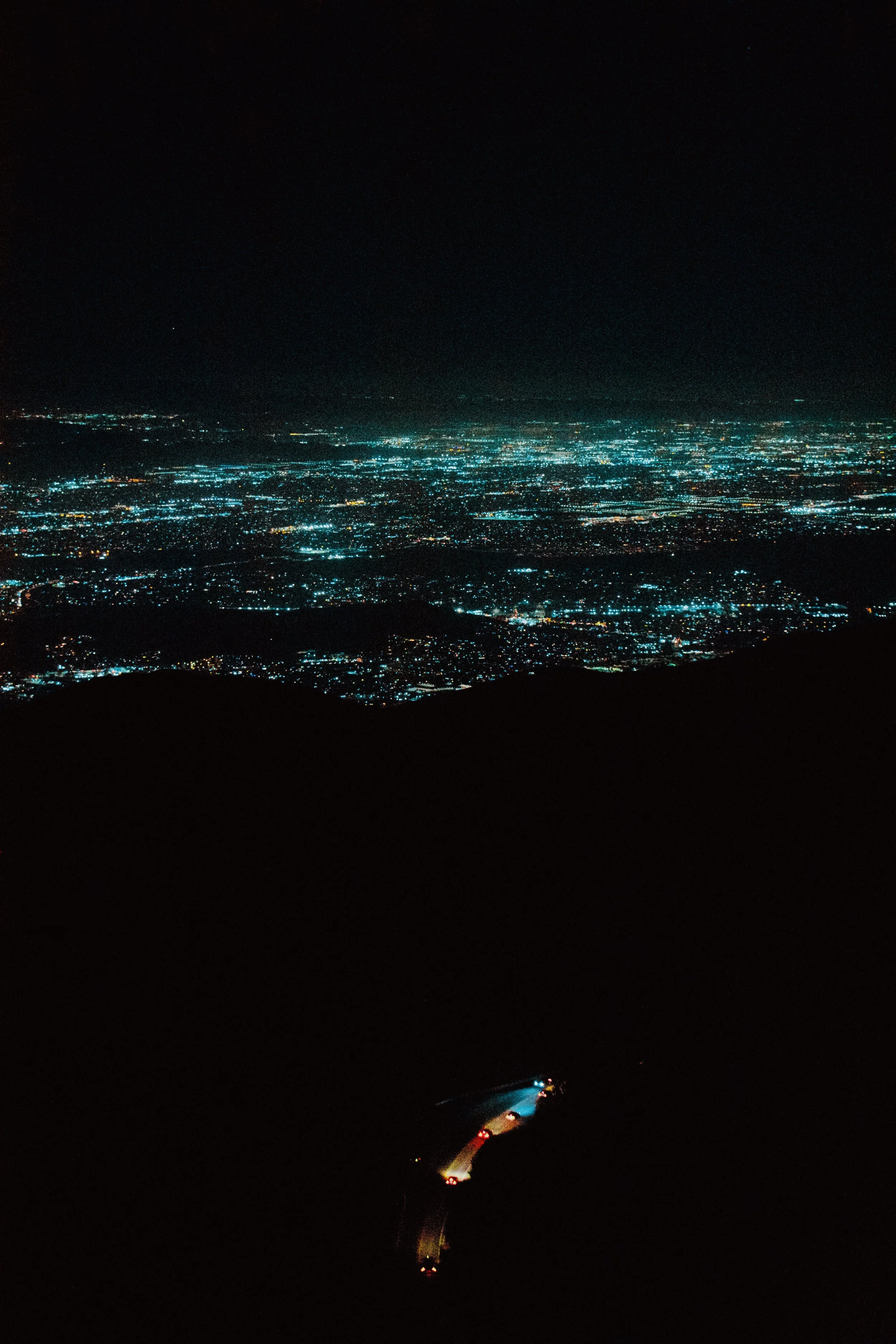 Nighttime aerial view of a distant city with illuminated streets and buildings, seen from above over dark mountains, with a faint trail of a vehicle on a nearby road.