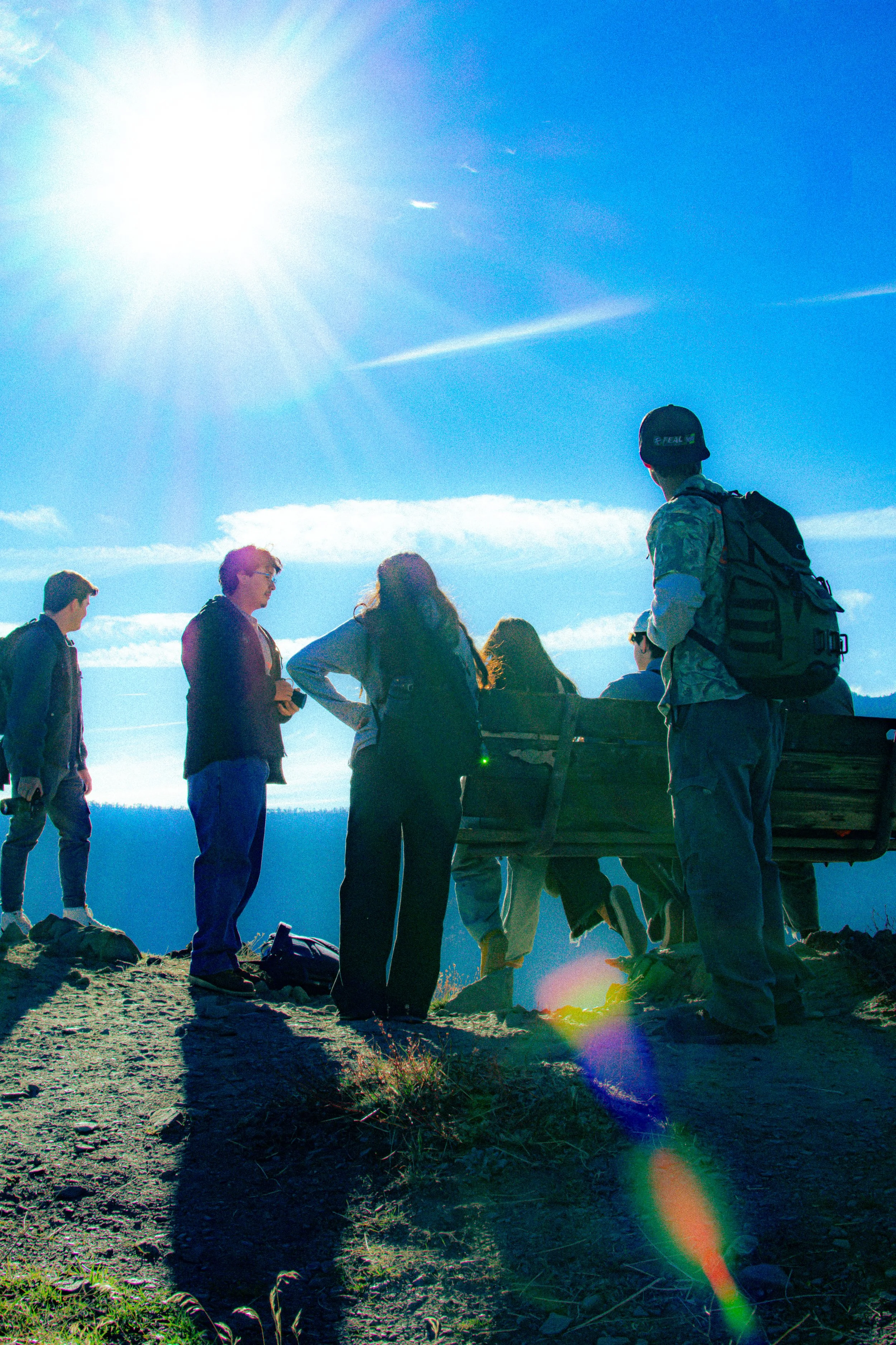 A group of people gathered outdoors on a sunny day, with some standing and others sitting on a wooden bench, enjoying the view.