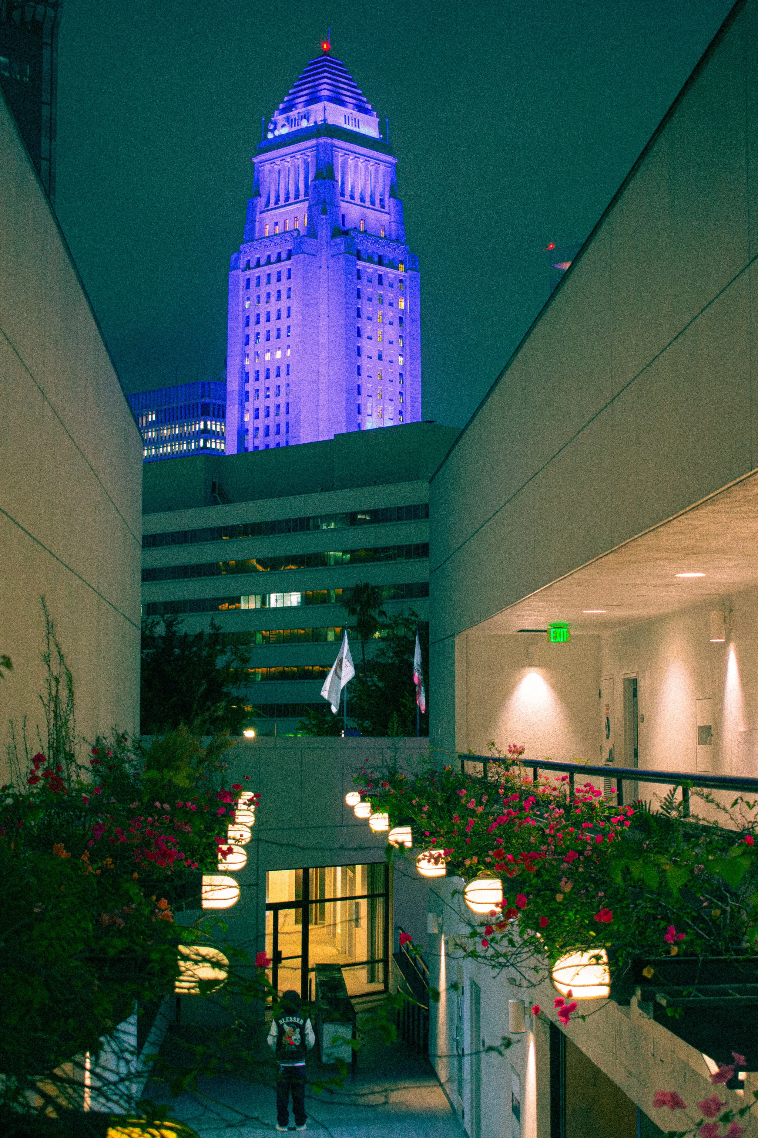 Night view of a tall, illuminated Los Angeles City Hall with a purple light, framed by modern white buildings, flower arrangements, and hanging lanterns at a rooftop or terrace.