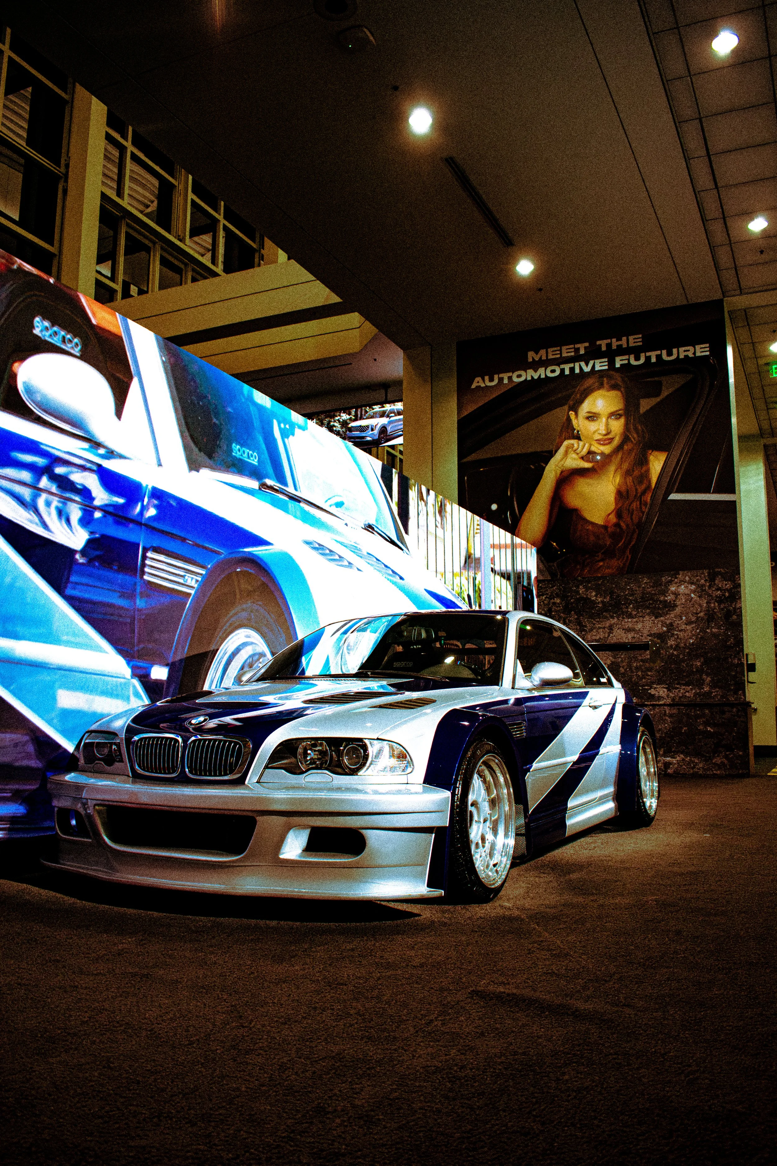 A silver and blue race car on display in front of large digital screens and a poster with a woman and the text 'Meet the Automotive Future'.